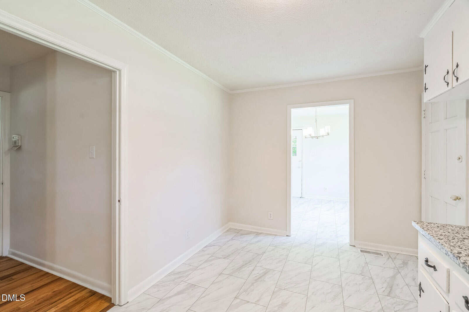 Bright, empty room with light tile floor and off-white walls, leading to another room with a chandelier and large window, visible through an open doorway. Part of a kitchen counter with granite can be seen on the right.