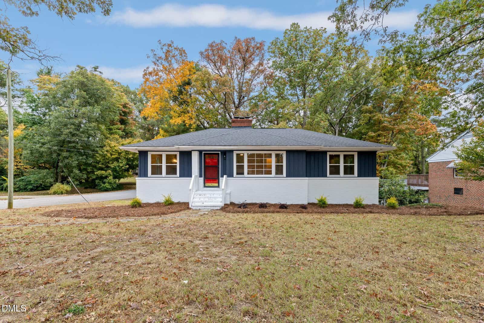 A single-story house at 1105 Dabney Drive, Henderson, features a white and dark blue exterior, a red front door, a small front porch with steps, and a lawn with sparse grass, surrounded by trees with autumn foliage.