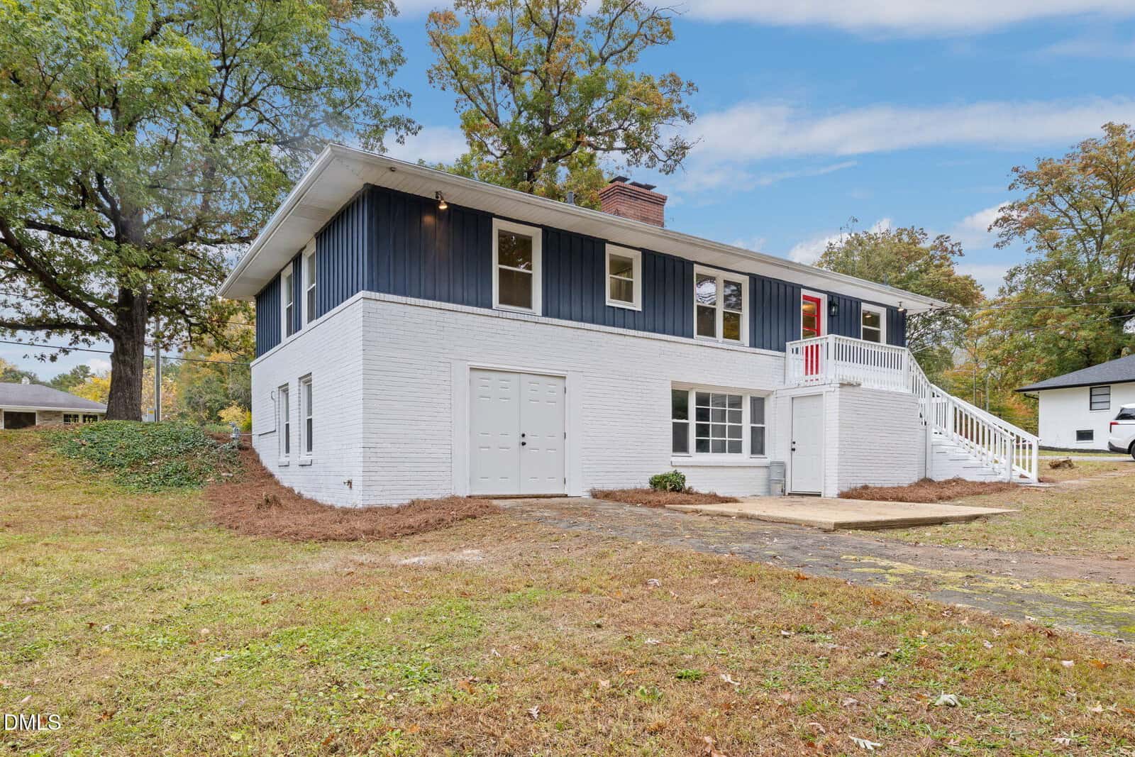 A two-story house with a white brick lower level and dark blue upper siding, a white staircase leading to a front entrance, and a yard with grass and trees in the background.
