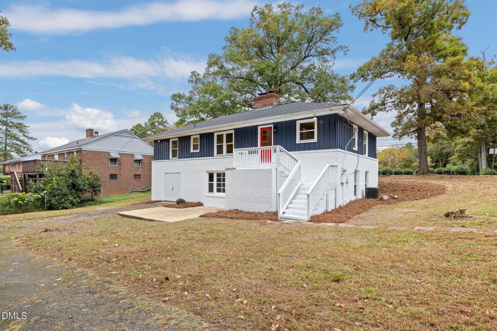 A two-story house with white brick on the lower level and dark blue siding on the upper level, white stairs, a red front door, and a spacious yard with trees and a driveway on the left.