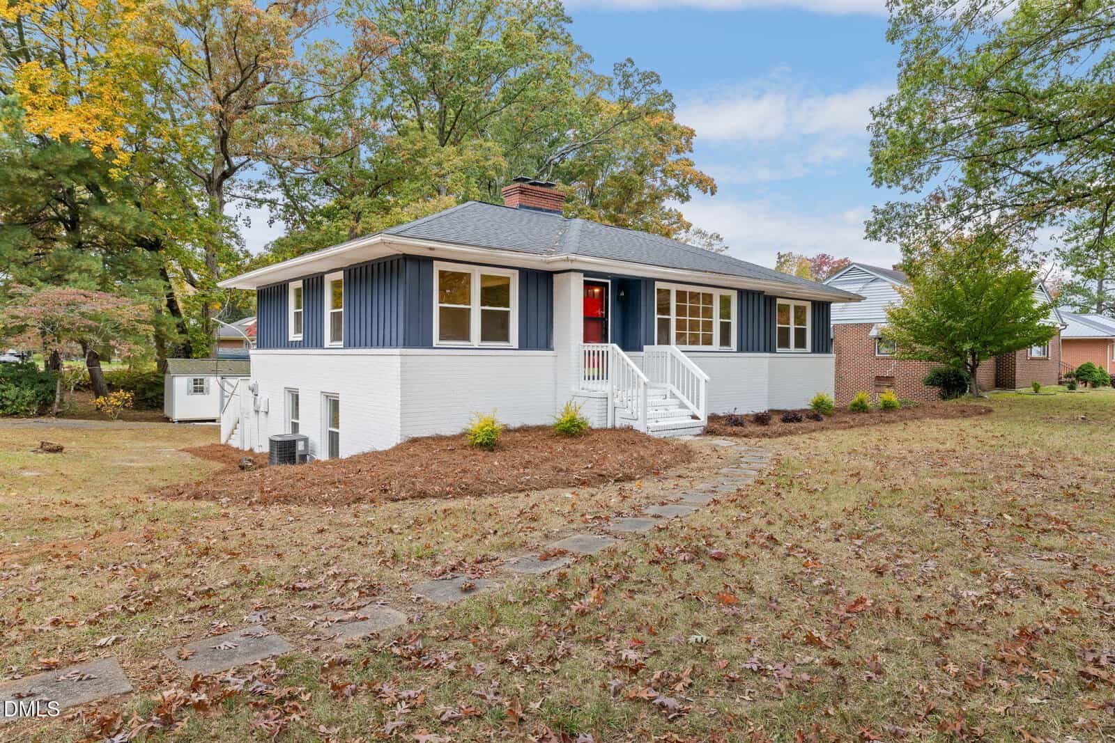 A single-story house with blue siding and white brick exterior, a gray shingled roof, a red front door, white railing, steps leading to the entrance, and a yard with trees and a stone pathway.
