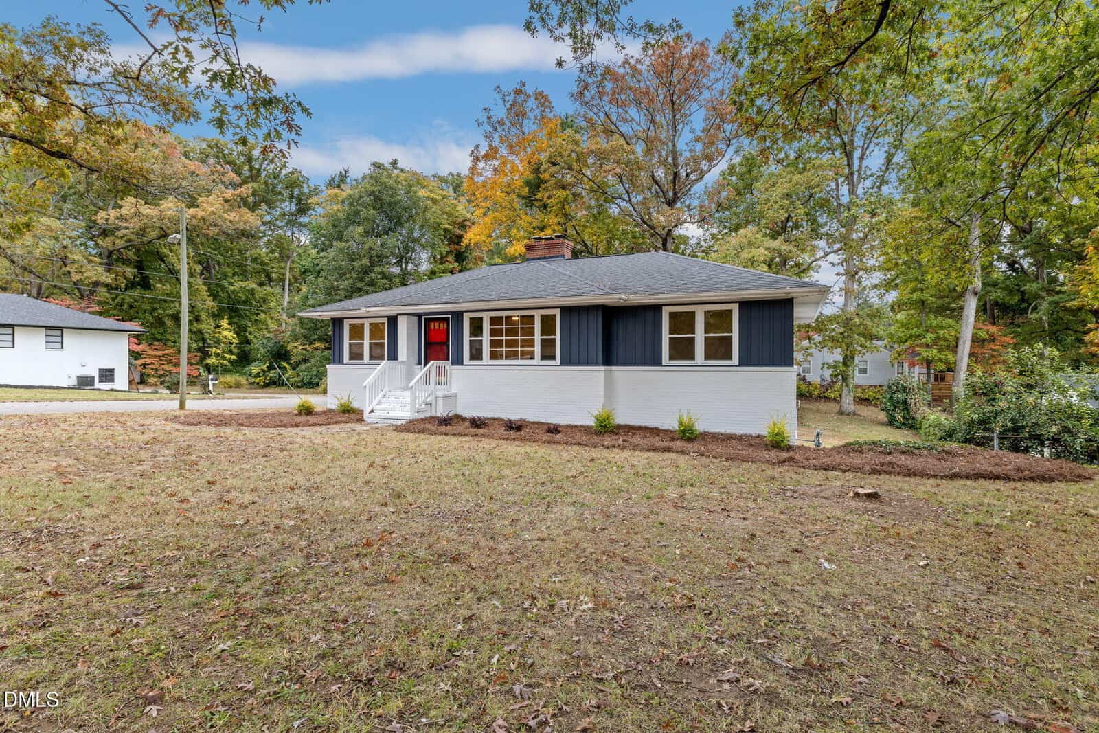 Single-story house with dark blue siding and white lower walls, a red front door, white steps, and small bushes planted along the front, surrounded by grass and mature trees under a partly cloudy sky.