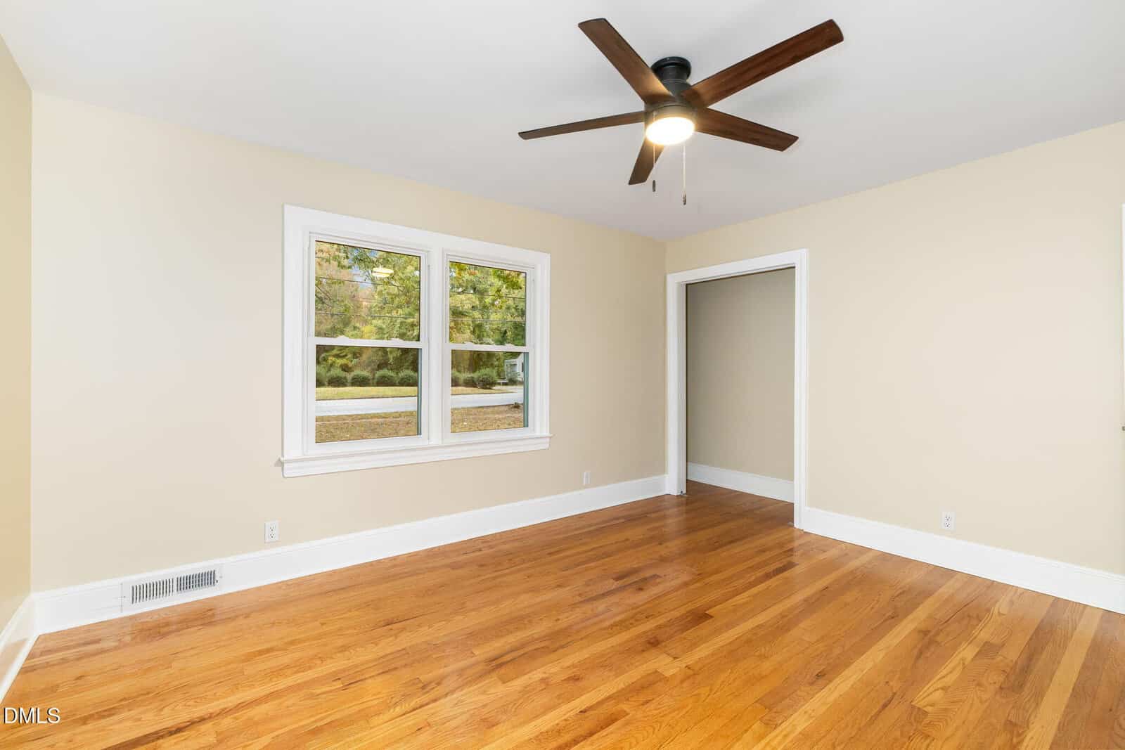 Empty room with light-colored walls, hardwood floors, a ceiling fan with a light, a double window, white trim, and an open doorway leading to another room.