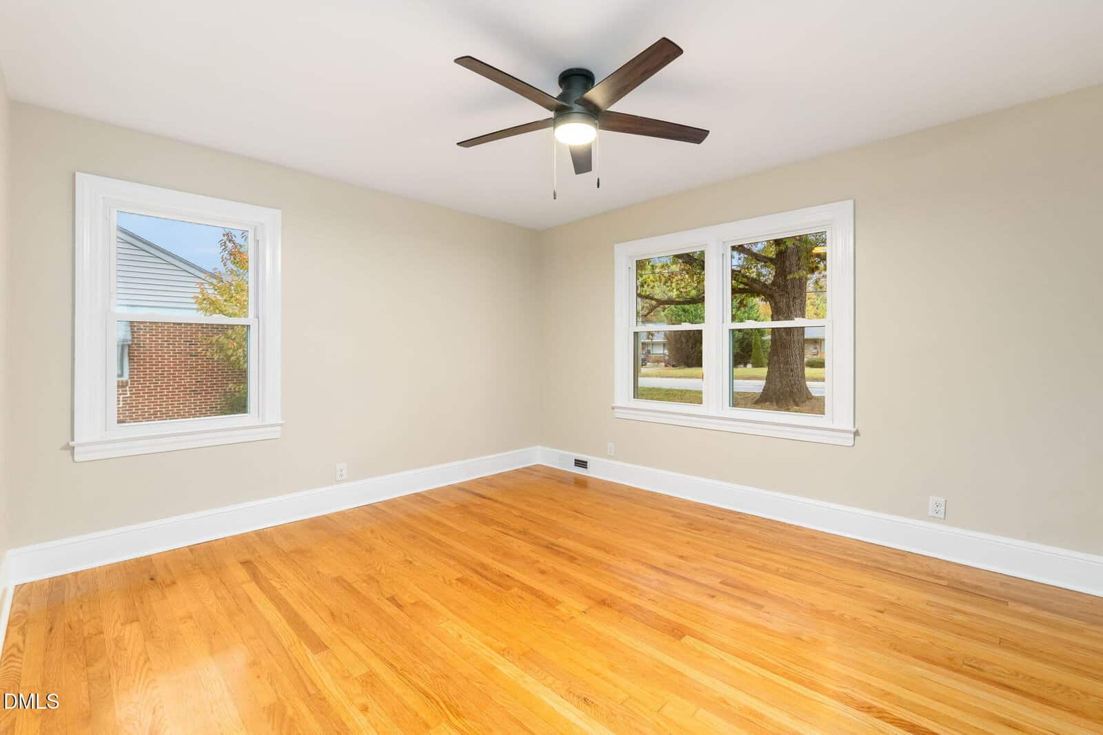 Empty room with light wood floor, beige walls, white trim, a ceiling fan with light, and two windows showing views of trees and a brick building outside.