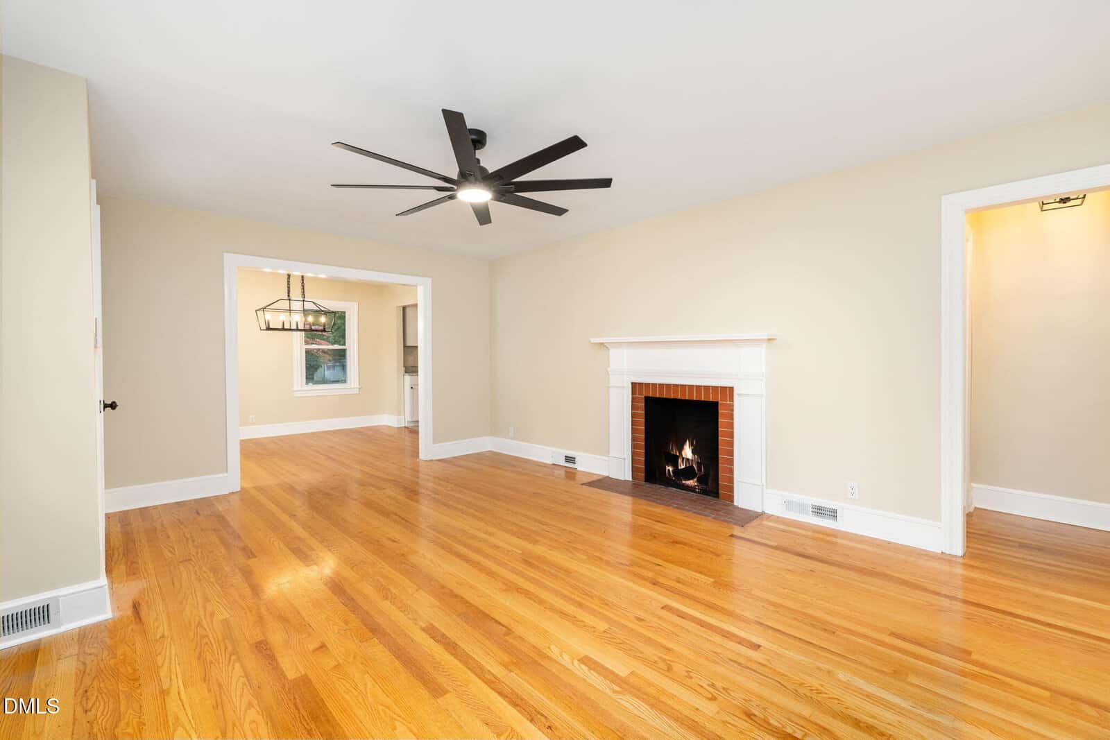 A spacious living room with polished hardwood floors, beige walls, a white fireplace with a lit fire, a modern ceiling fan, and white trim around doorways and baseboards. An adjoining room is visible in the background.