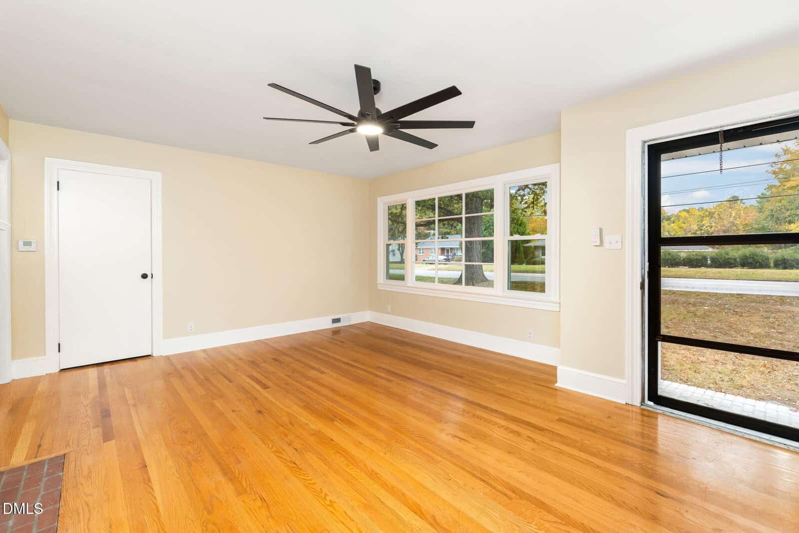 A bright, empty room with light tan walls, hardwood floors, a large black ceiling fan, a white door, a set of three windows, and a glass door leading outside.