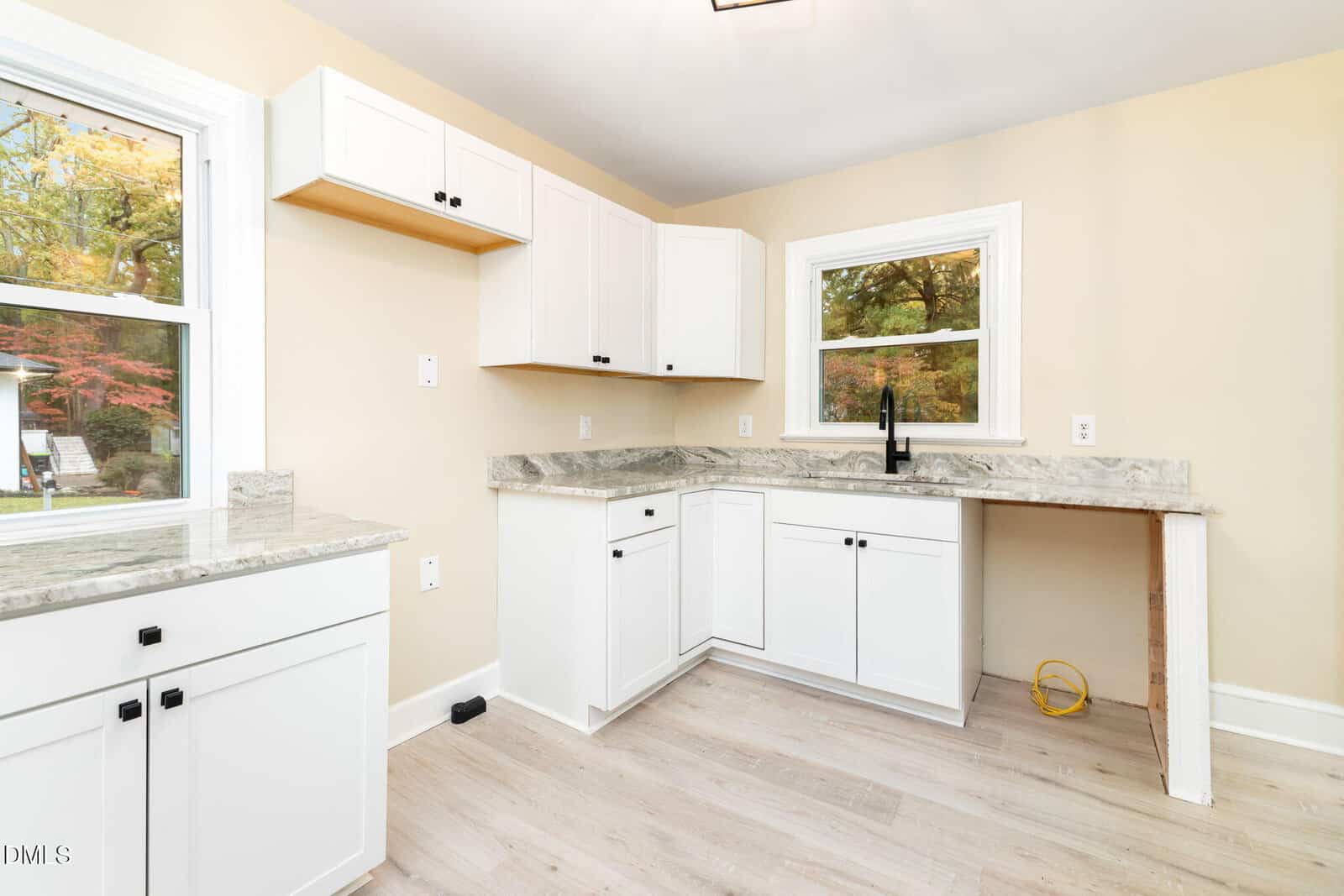 A kitchen with light-colored walls, white cabinets, marble countertops, light wood flooring, and two windows. The sink is installed, but there are no appliances present. Some cabinet doors are open.