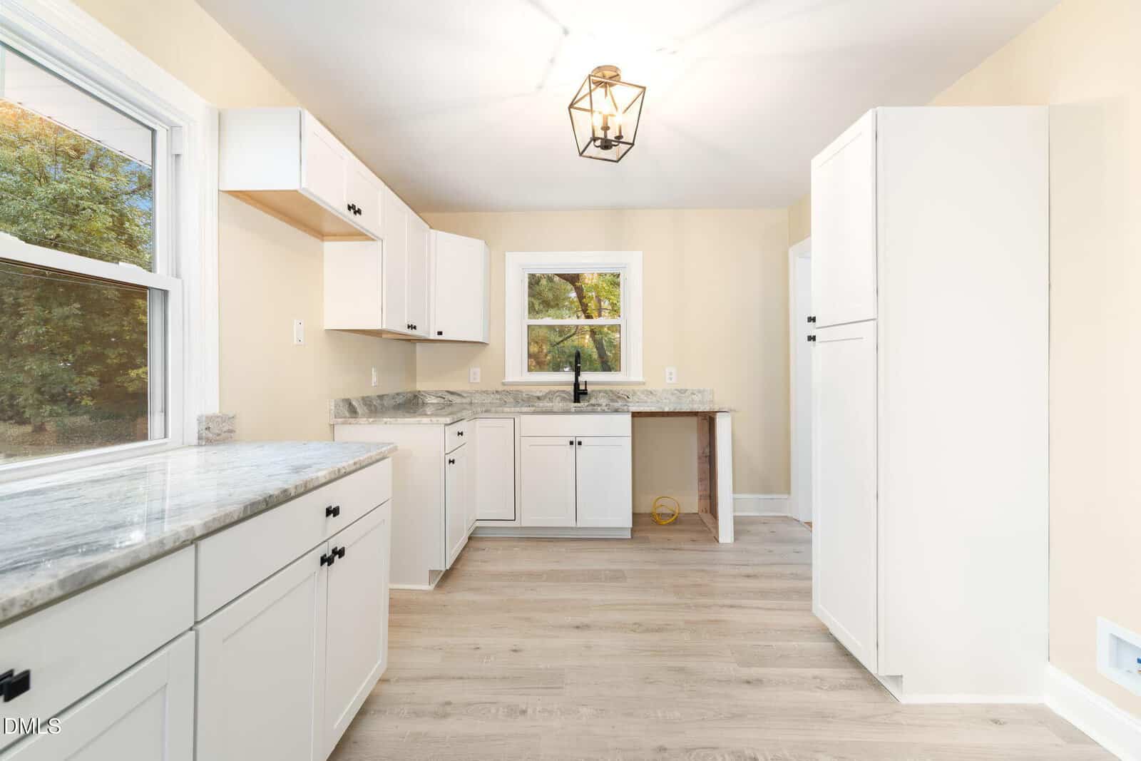 A kitchen with white cabinets, light wood flooring, marble countertops, and a window above the sink. There is a modern light fixture on the ceiling and trees are visible outside the window and left-side door.