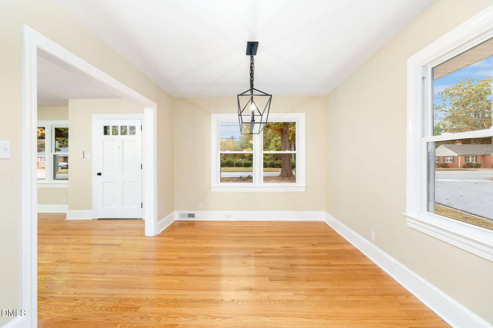A bright, empty room with light beige walls, hardwood floors, two windows, a geometric black pendant light, and an open doorway leading to another room with a white front door.