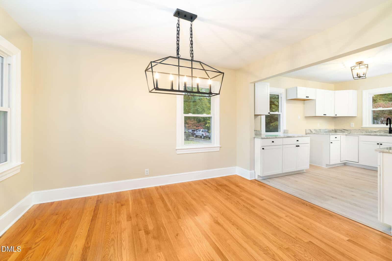 Bright dining area with hardwood floors, a modern geometric chandelier, and an open view into a white kitchen with large windows, light countertops, and wood-style flooring.