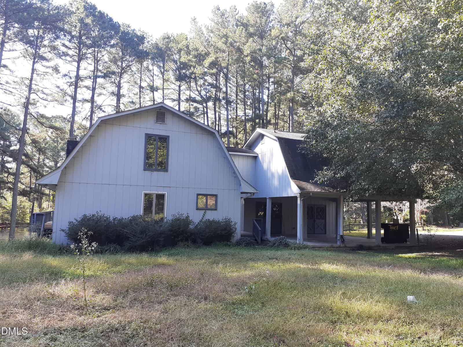 A two-story house at 1576 Munns Road, Creedmoor, features gray siding, a barn-style roof on the left wing, and a covered porch on the right, surrounded by tall pine trees and overgrown grass in the yard.