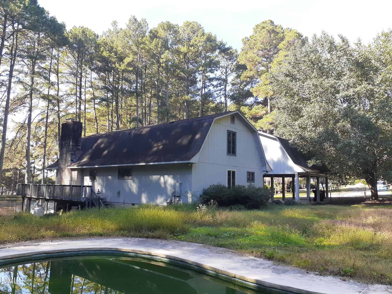 A two-story, barn-style house with gray siding and a black roof is surrounded by tall pine trees. There is a covered carport area and an in-ground pool with green water in the foreground.