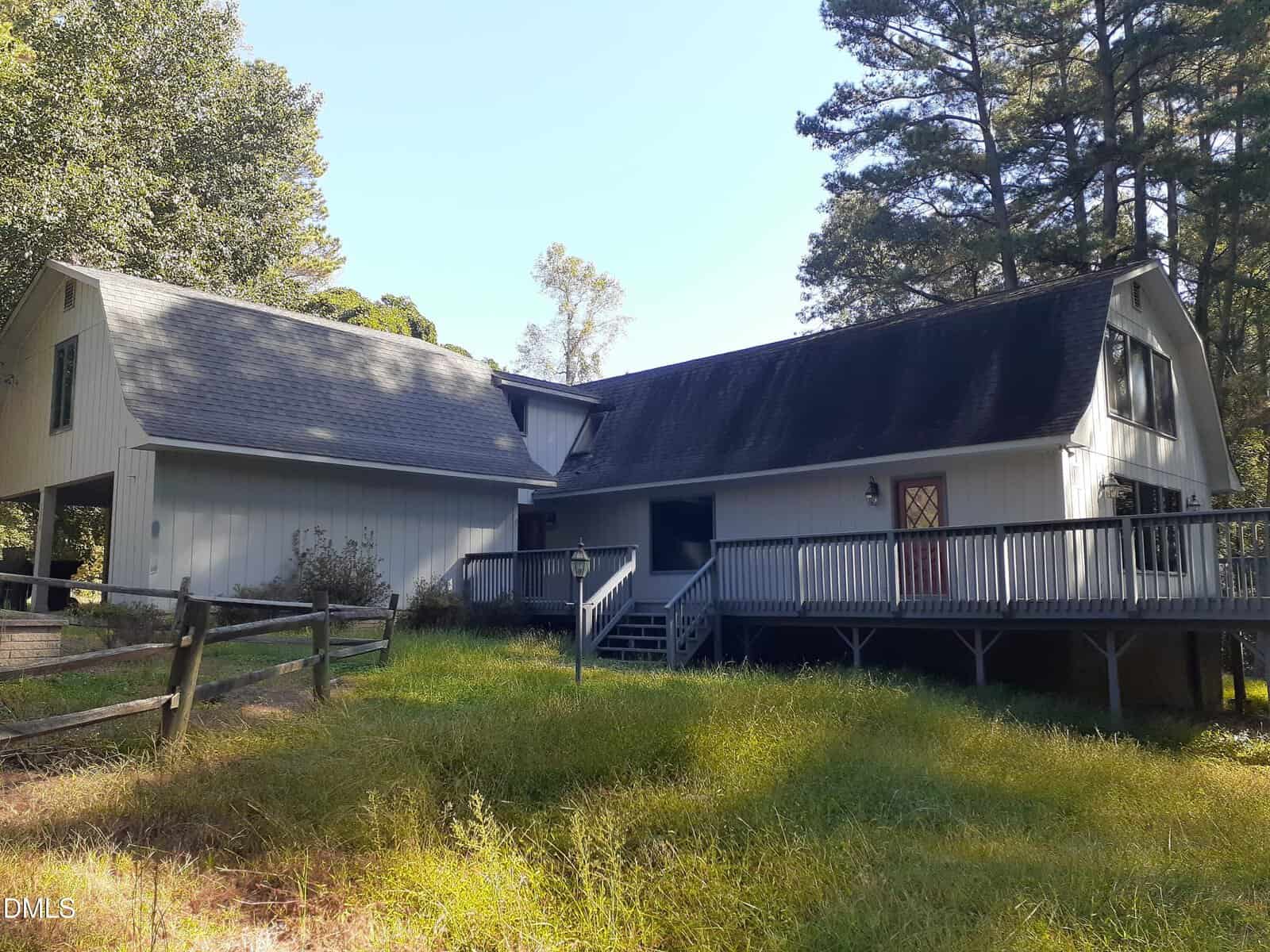 A two-story gray house with a sloped roof, large wraparound porch, wooden ramp, and steps. The yard has tall grass, a split-rail fence, and is surrounded by trees under a clear sky.
