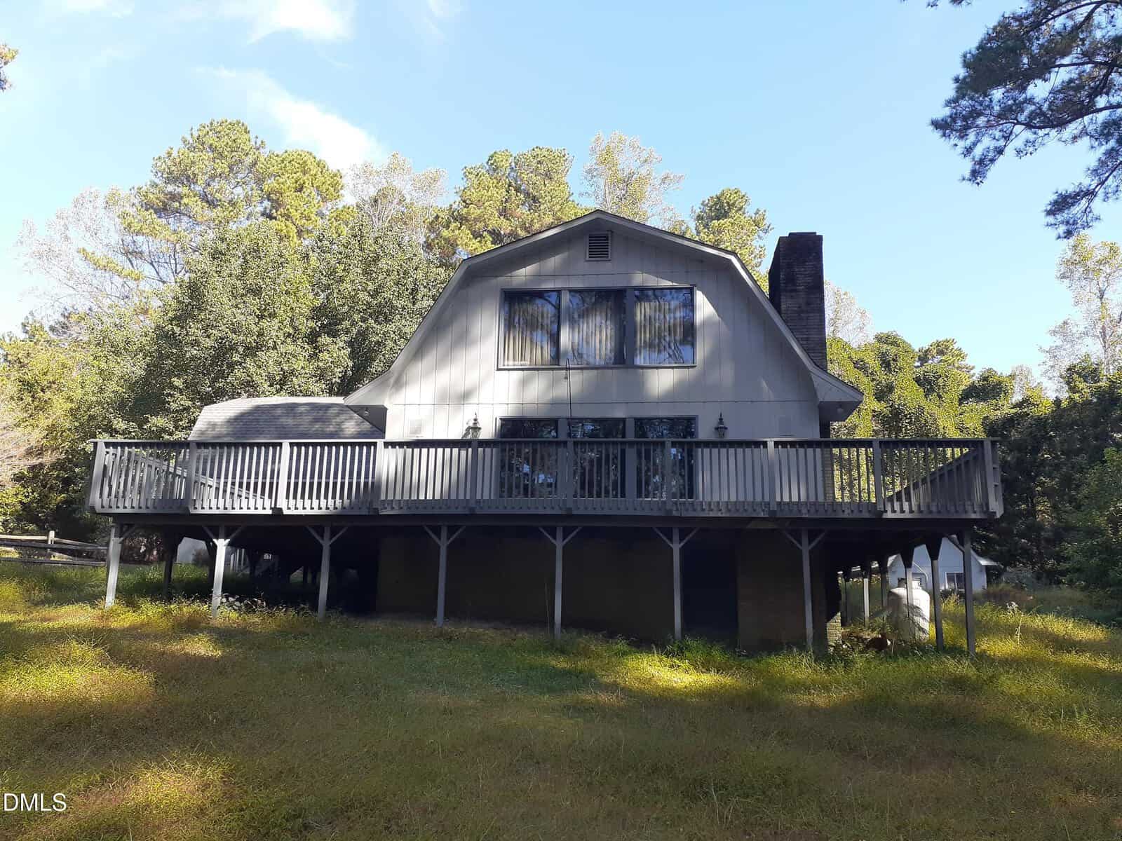 A two-story house with a gray exterior and a large wraparound wooden deck, elevated on stilts, set among trees and grass under a clear blue sky.
