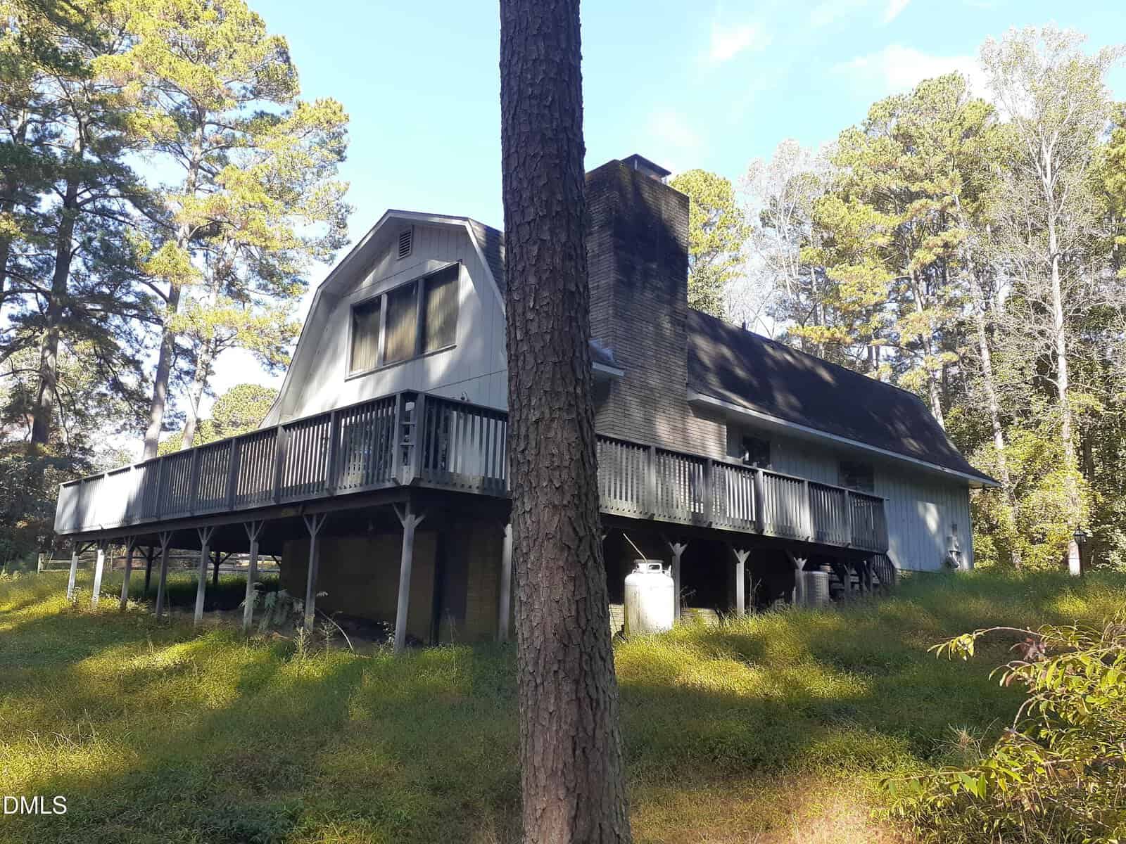 A two-story house with a gambrel roof and elevated wooden deck sits among tall trees and grass, with a large chimney on one side and a propane tank visible underneath the deck.