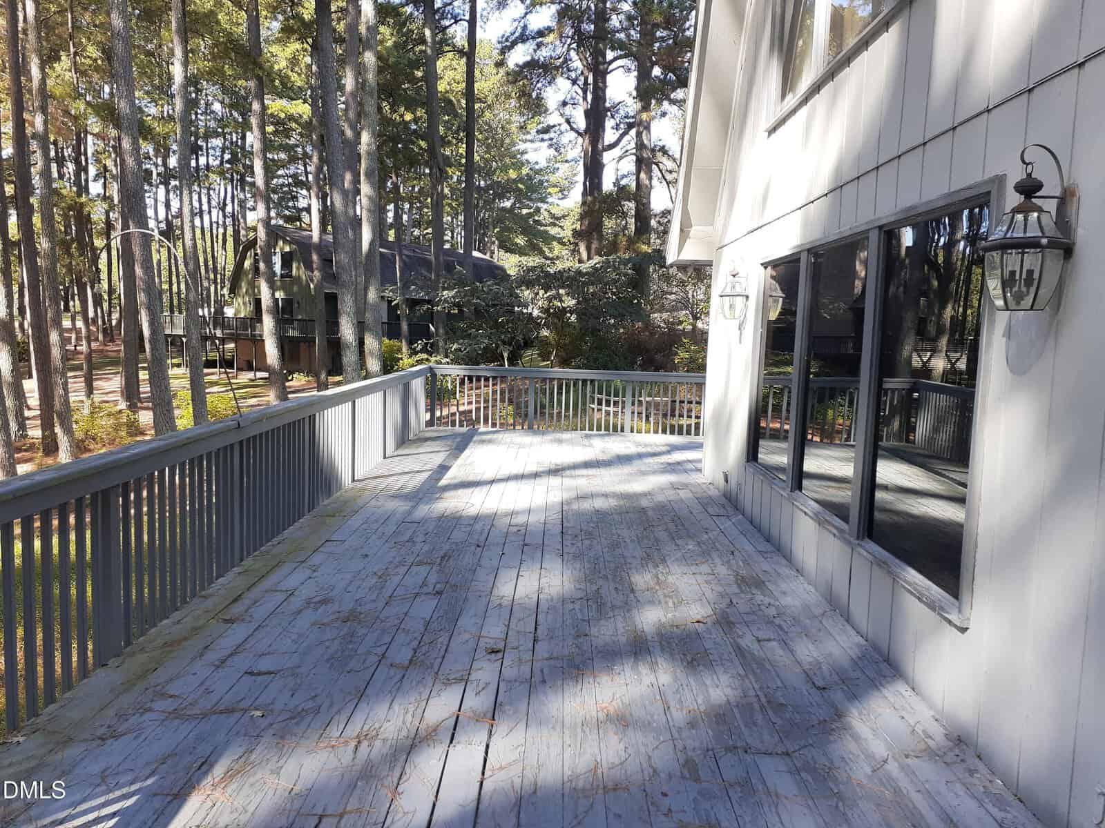 A large wooden deck with weathered blue-gray paint is attached to a house with gray siding and large windows. The deck overlooks a wooded area with tall pine trees and nearby houses.