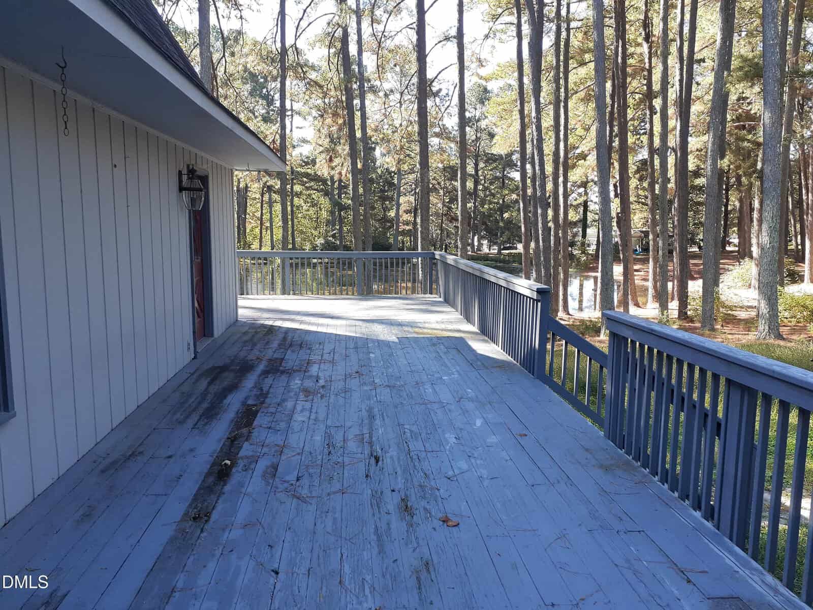A large wooden deck with worn blue-gray paint extends from a house, bordered by a simple railing and overlooking a yard with many tall pine trees. Sunlight filters through the trees, casting shadows on the deck.