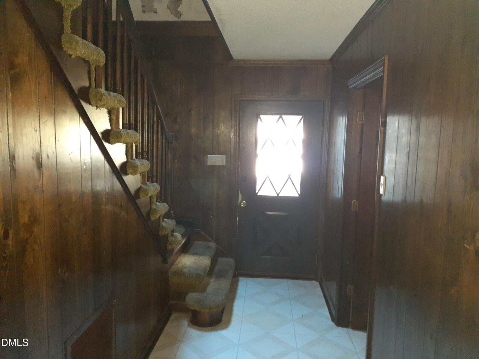 Interior hallway with dark wood-paneled walls, a staircase with carpeted steps and railing on the left, a door with a diamond-patterned window in the center, and tiled floors. Light enters through the door window.