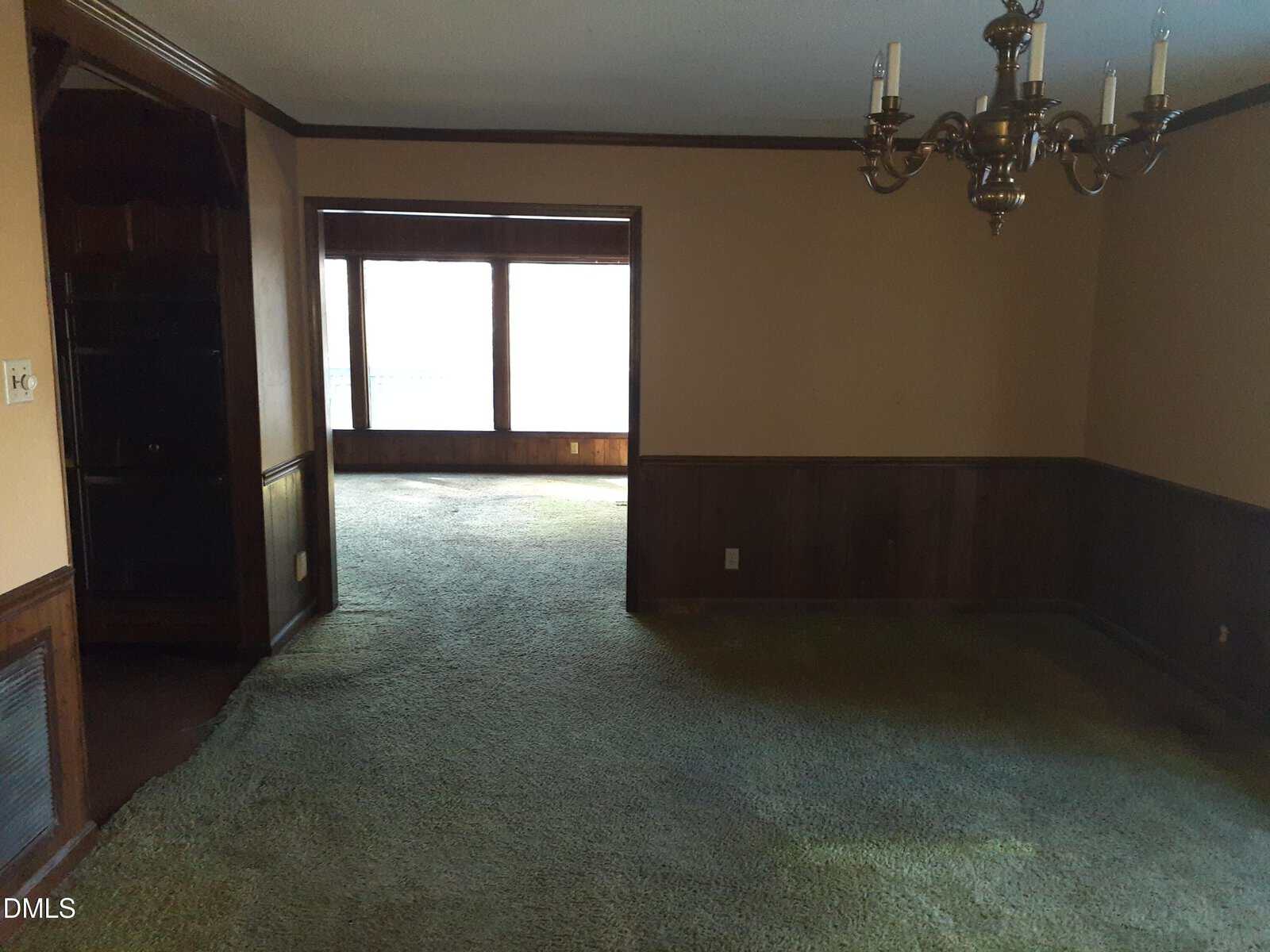 A dimly lit room with green carpet, wood-paneled walls, a brass chandelier, and a large open entryway leading to a sunlit area with big windows.