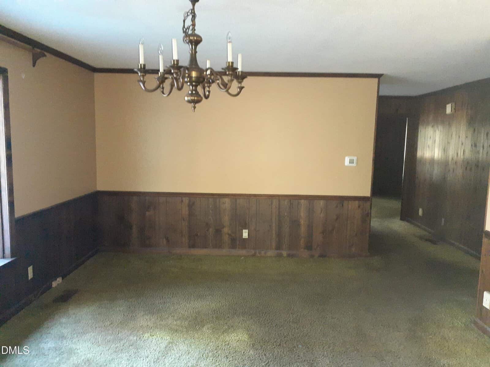 A room with wood-paneled walls, green carpet, a brass chandelier, beige upper walls, and an open doorway leading to a hallway. Natural light enters from a window on the left.