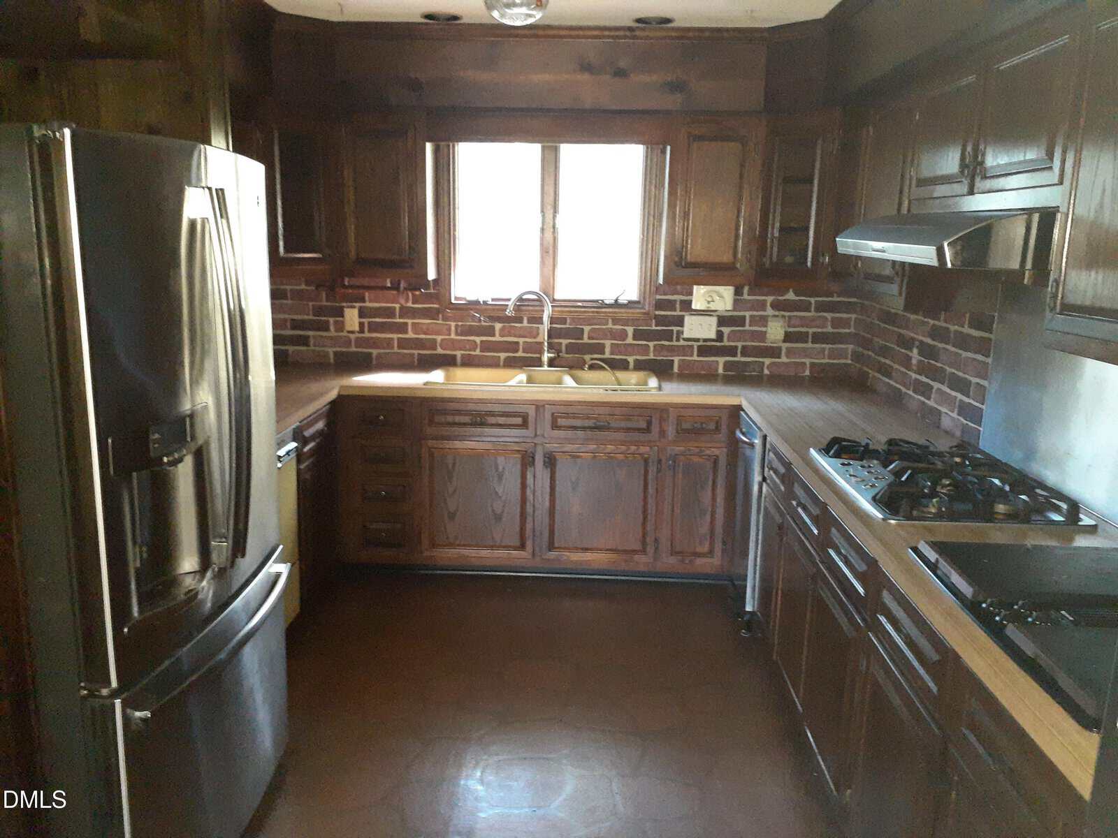 A kitchen with dark wood cabinets, brick backsplash, stainless steel fridge, gas stove, and a double sink under a window. The counters are light brown, and the floor is a reddish-brown tile.