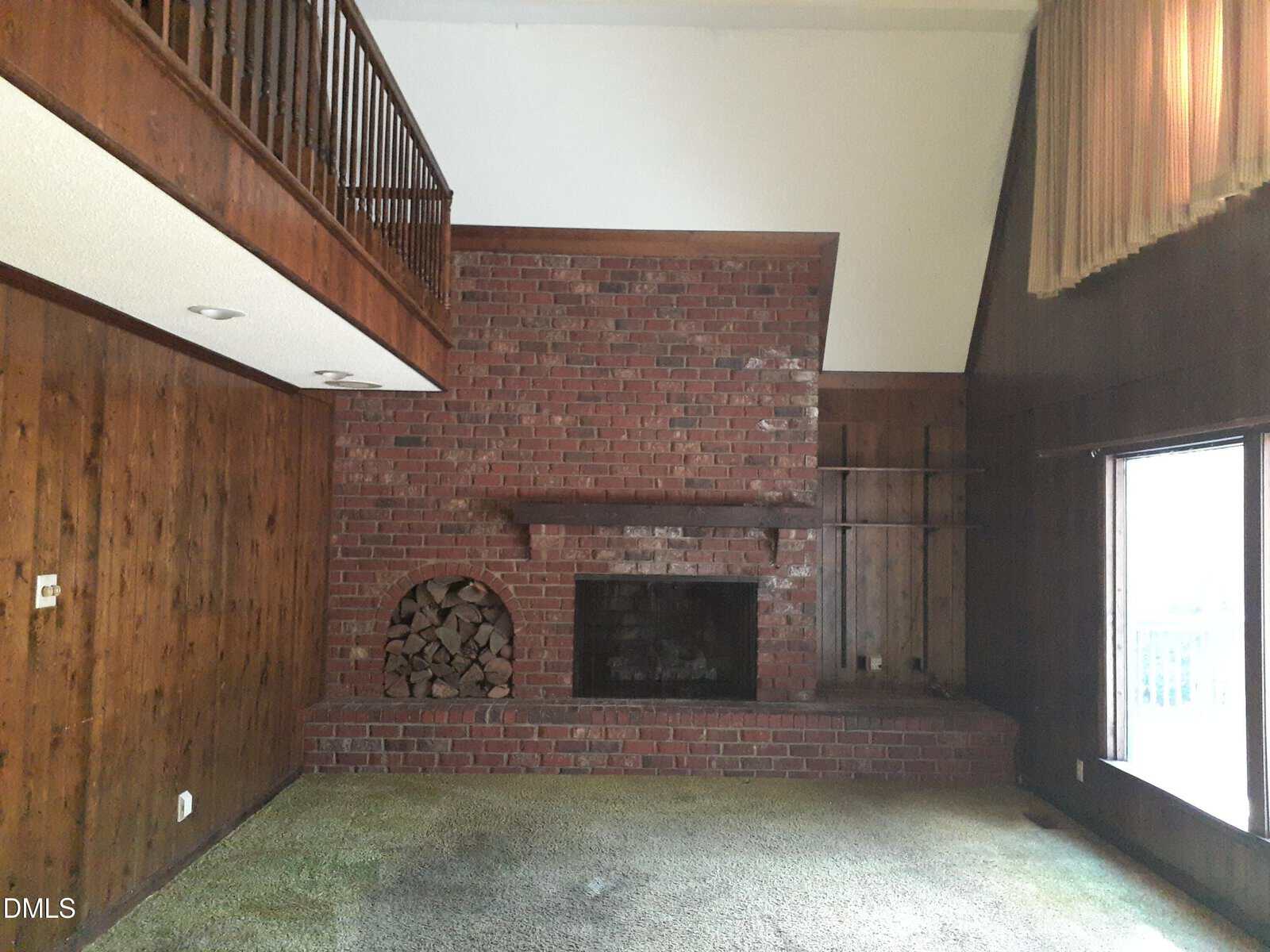 A living room with dark wood-paneled walls, a brick fireplace with a log storage area, green carpet, a large window, and a wooden balcony above. Curtains hang on the right side, and the ceiling is vaulted.