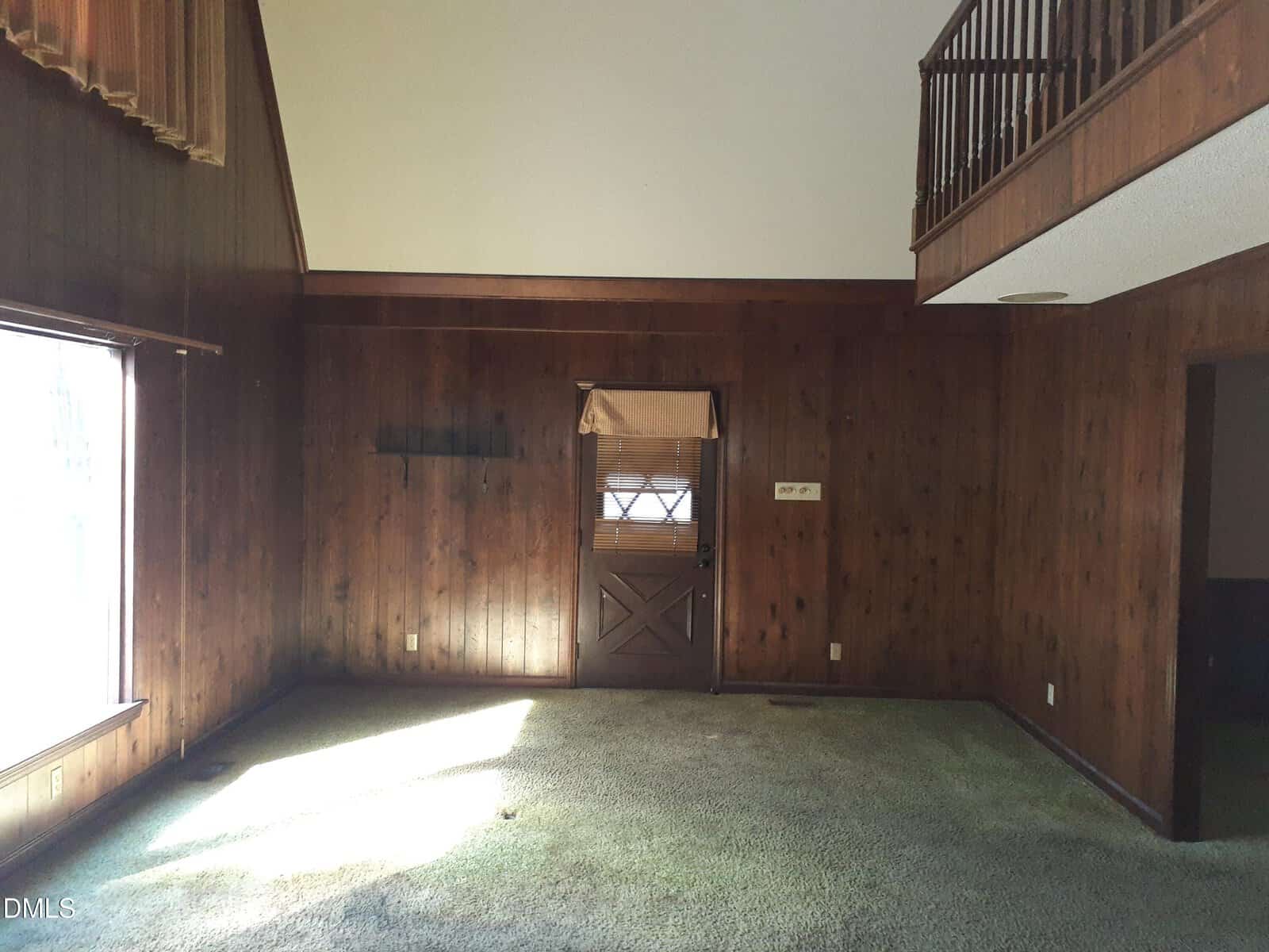 A room with dark wood-paneled walls, greenish carpet, a window letting in sunlight, a door with a small window and blinds, and an upper balcony railing on the right above part of the room.