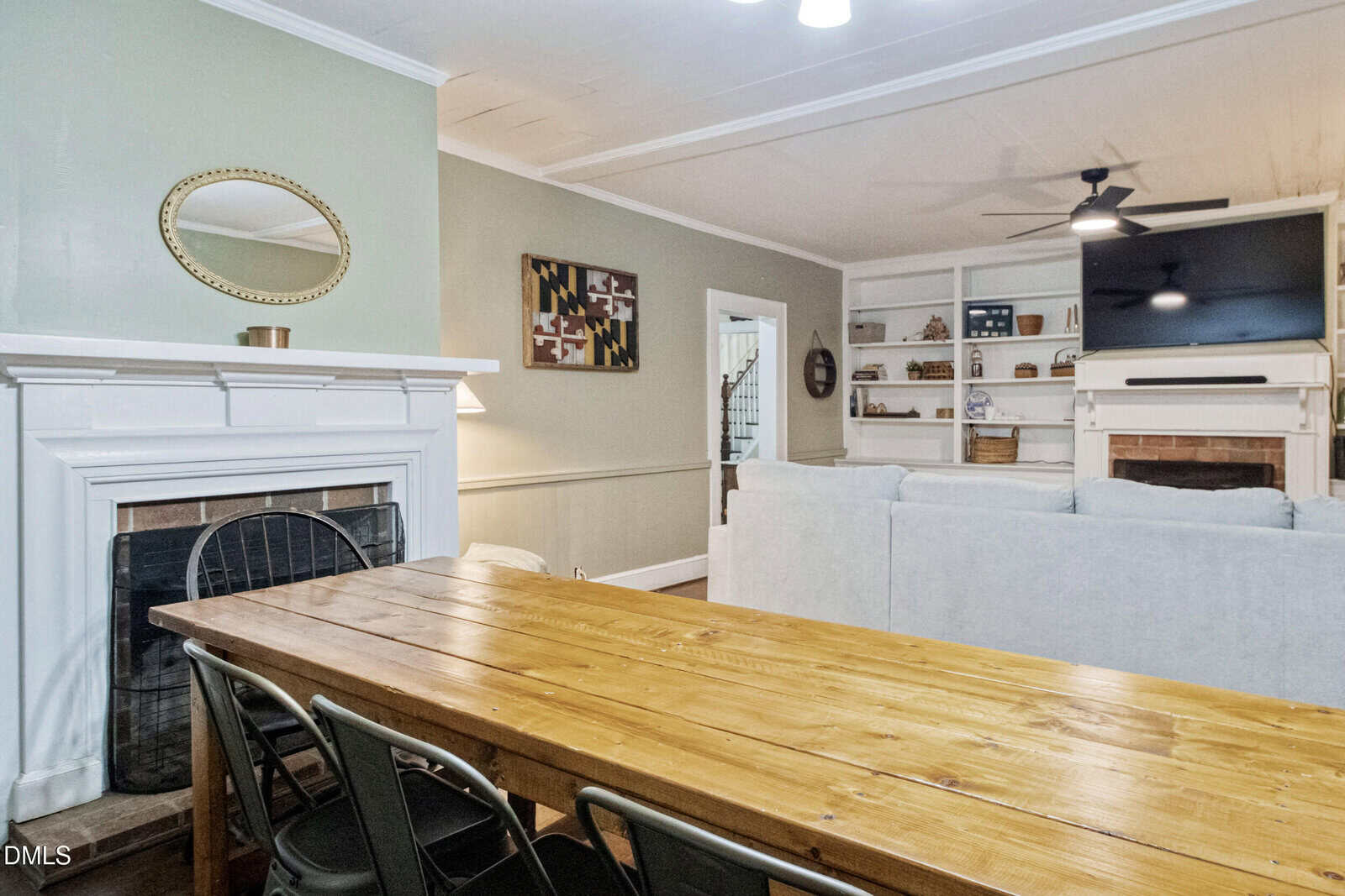A dining area with a wooden table and metal chairs is adjacent to a living area with a light-colored sofa, built-in shelves, a wall-mounted TV, a ceiling fan, and a fireplace with a round mirror above it.