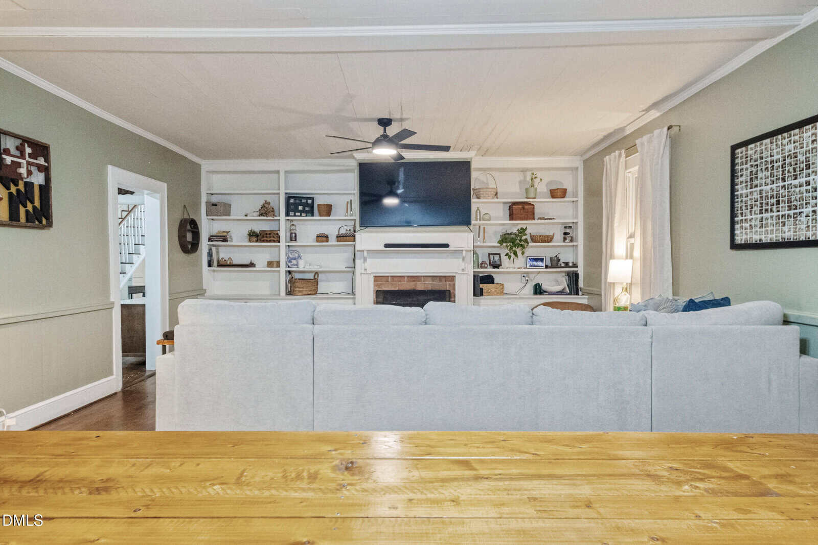 A living room with a light gray sectional sofa, a wall-mounted TV above a fireplace, built-in shelves with decor and baskets, and a wooden table in the foreground. White curtains cover windows on the right.