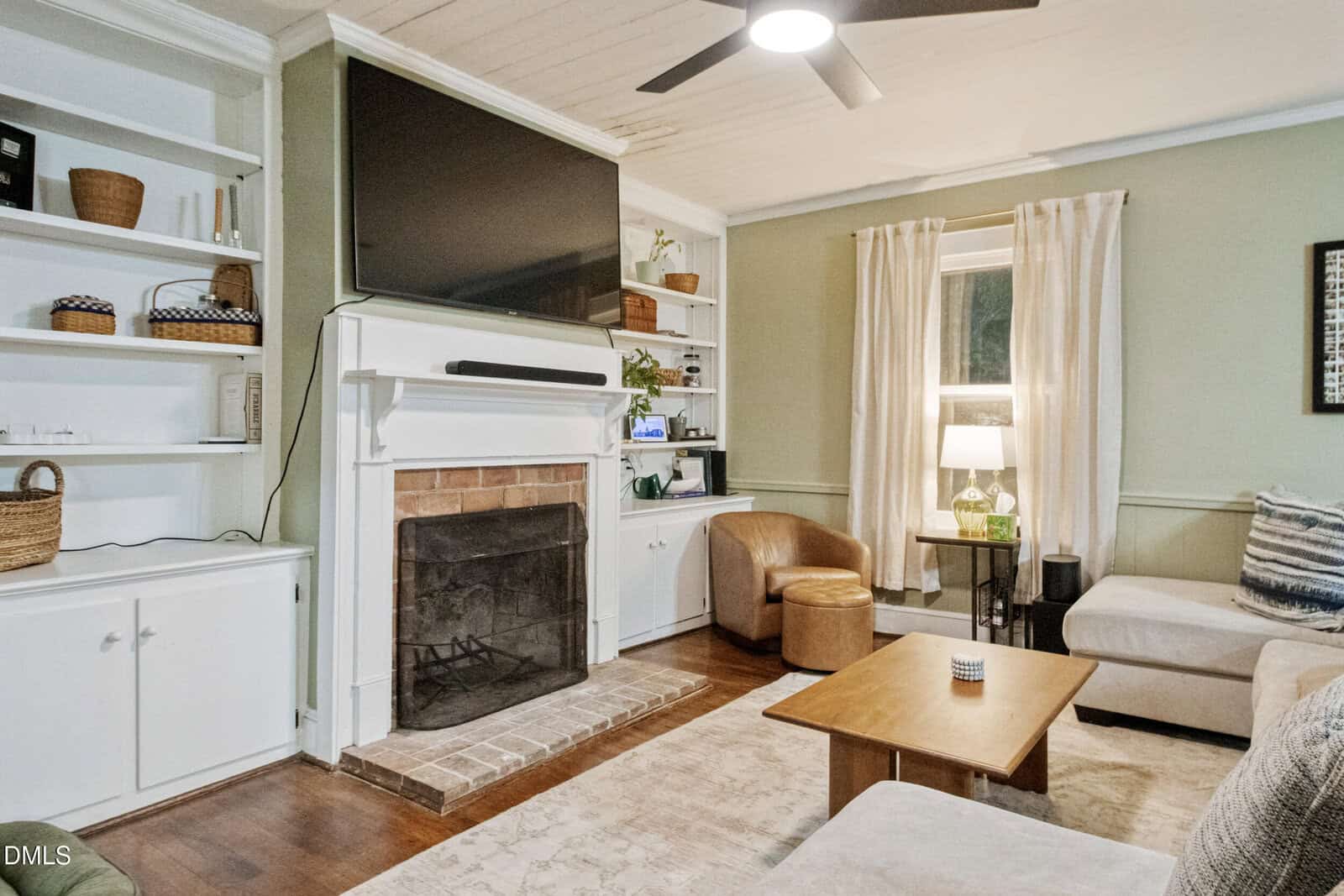 A living room with a wall-mounted TV above a white fireplace, built-in shelves on both sides, a sectional sofa, a wooden coffee table, a tan leather chair, and a window with cream curtains.