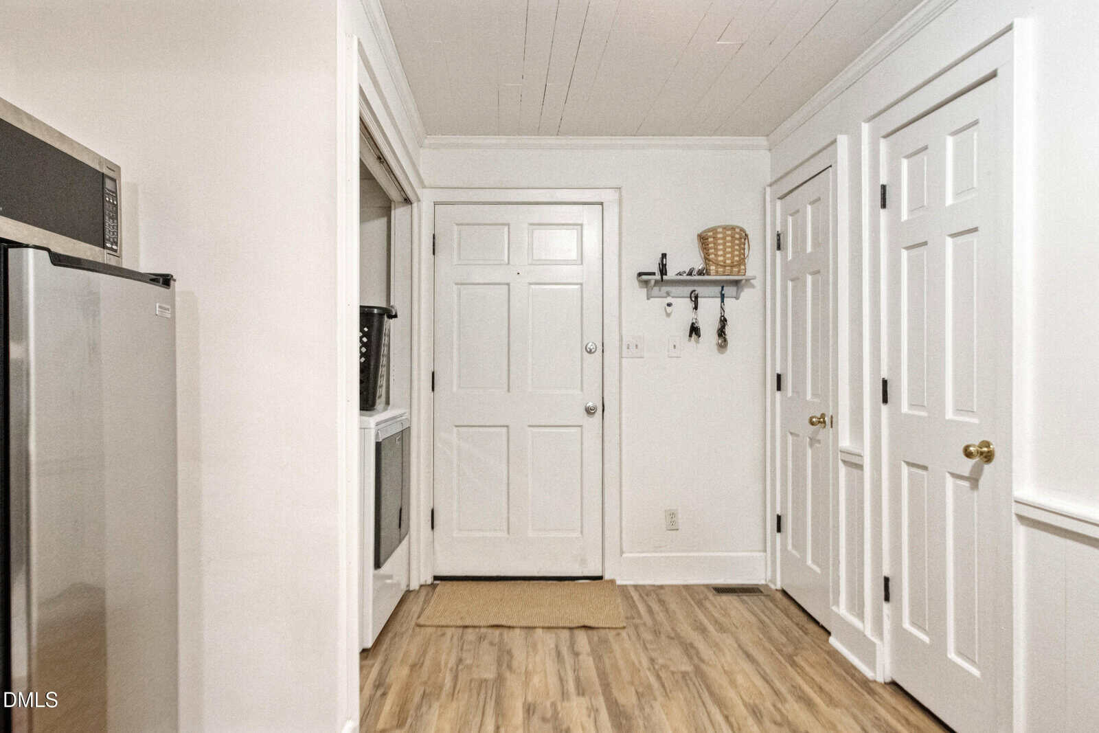 A white entryway with a closed door, three white closet doors, a small wall-mounted shelf with hooks holding a basket and keys, a beige rug, and wood-look flooring. Part of a stainless steel appliance is visible on the left.