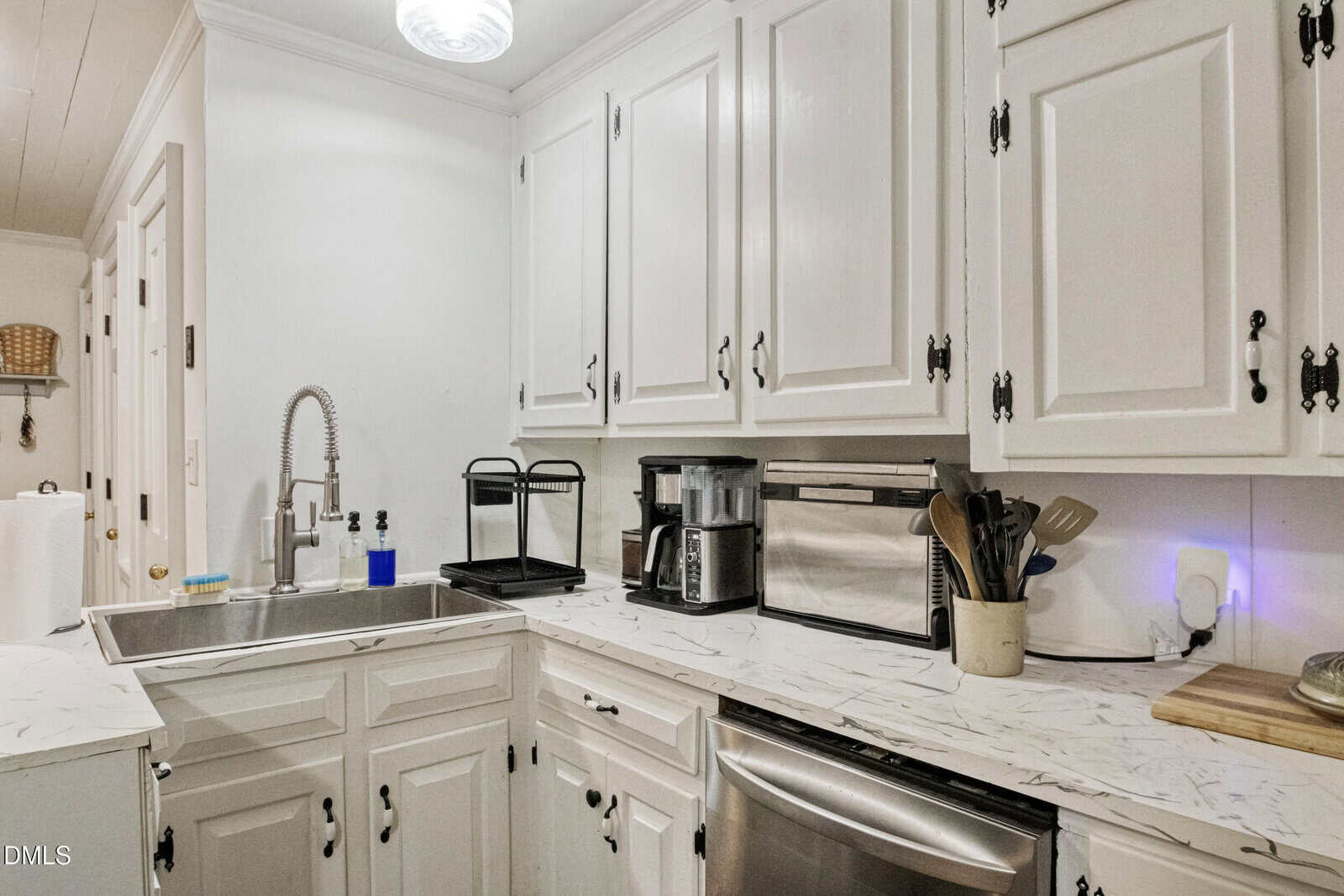 A white kitchen features a deep sink with a tall faucet, appliances like a toaster and coffee maker, utensils in a holder, and marble-patterned countertops beneath white cabinets with black hardware.