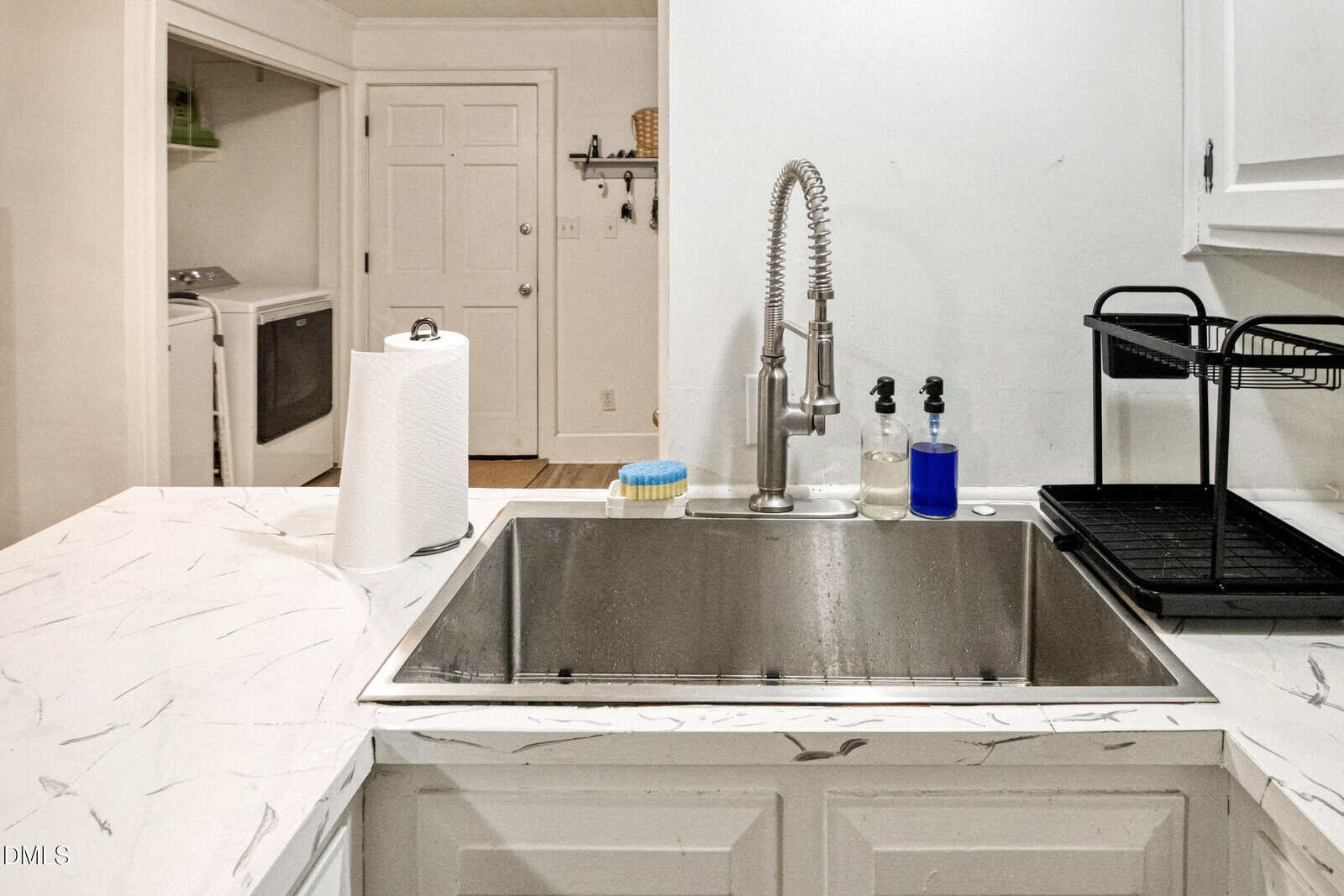 A stainless steel kitchen sink with a modern faucet, a roll of paper towels, soap dispensers, a dish brush, and a black dish rack on a white marbled countertop; a laundry area is visible in the background.