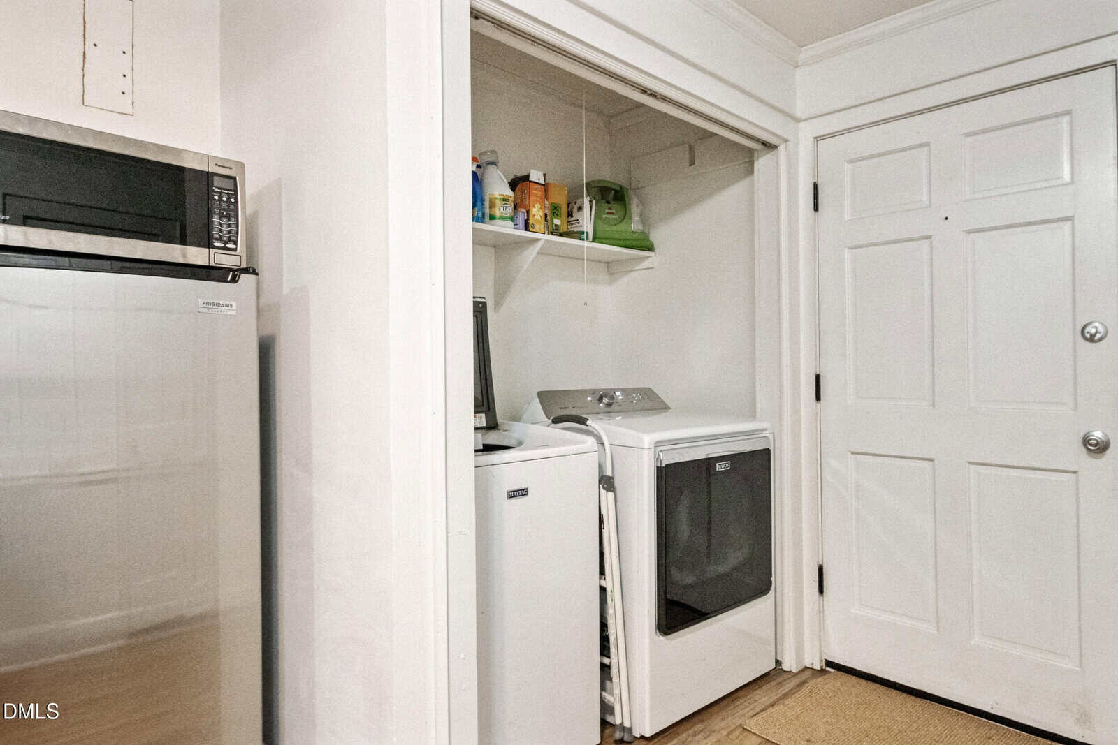 A small laundry area with a washing machine and dryer is set in a closet next to a white door. Shelves above hold detergent and other items. Part of a stainless steel refrigerator and microwave are visible on the left.
