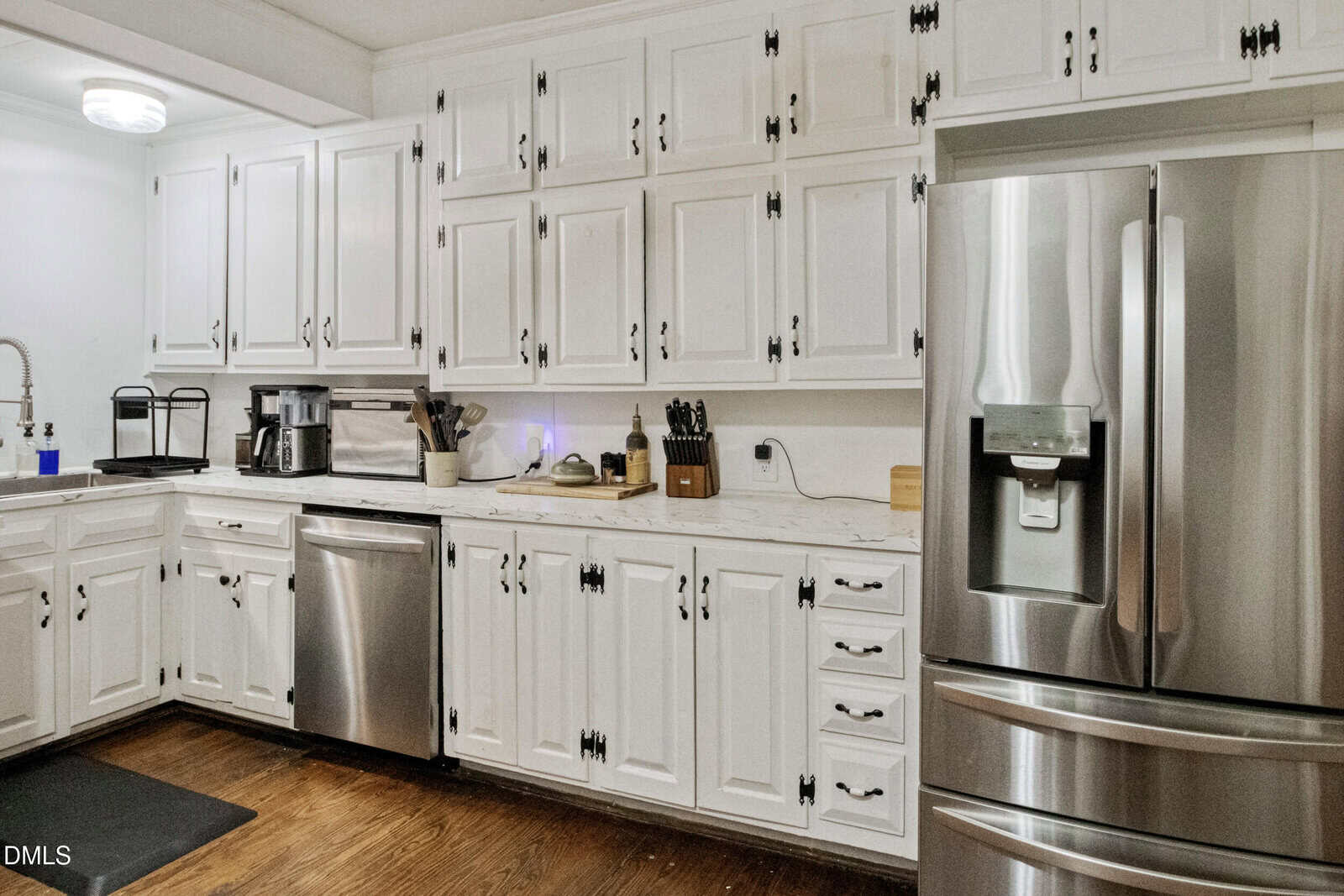 A modern kitchen with white cabinets, stainless steel appliances including a fridge and dishwasher, a coffee maker, knife set, and wooden floors.