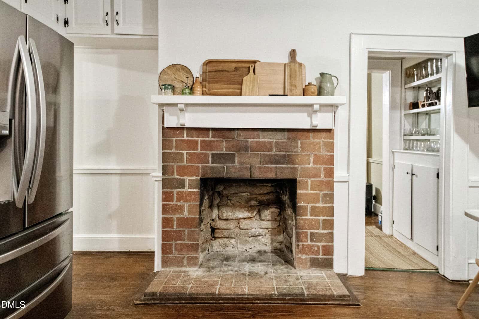 A brick fireplace with a white mantle, topped with cutting boards, dishes, and a pitcher, is set between white cabinets in a kitchen with wood floors and a stainless steel refrigerator nearby.
