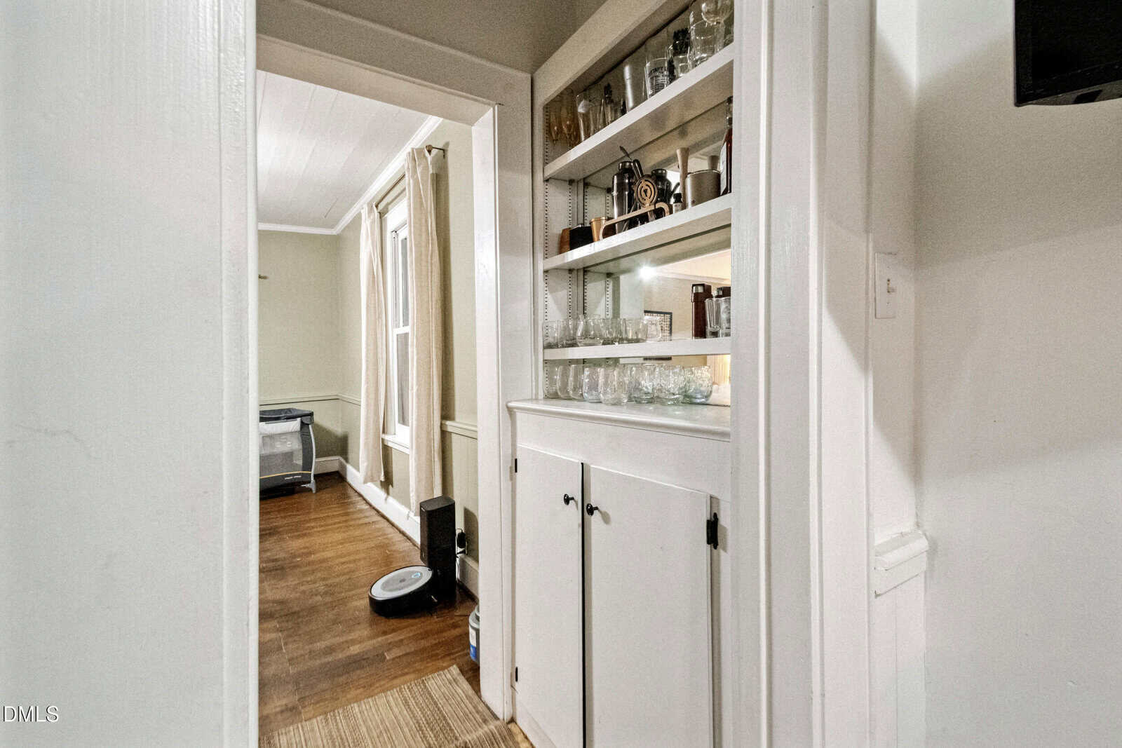 A small built-in white cabinet with shelves holding glassware and bottles is seen next to a doorway leading to a room with wood floors, a rug, and a robot vacuum near the wall.