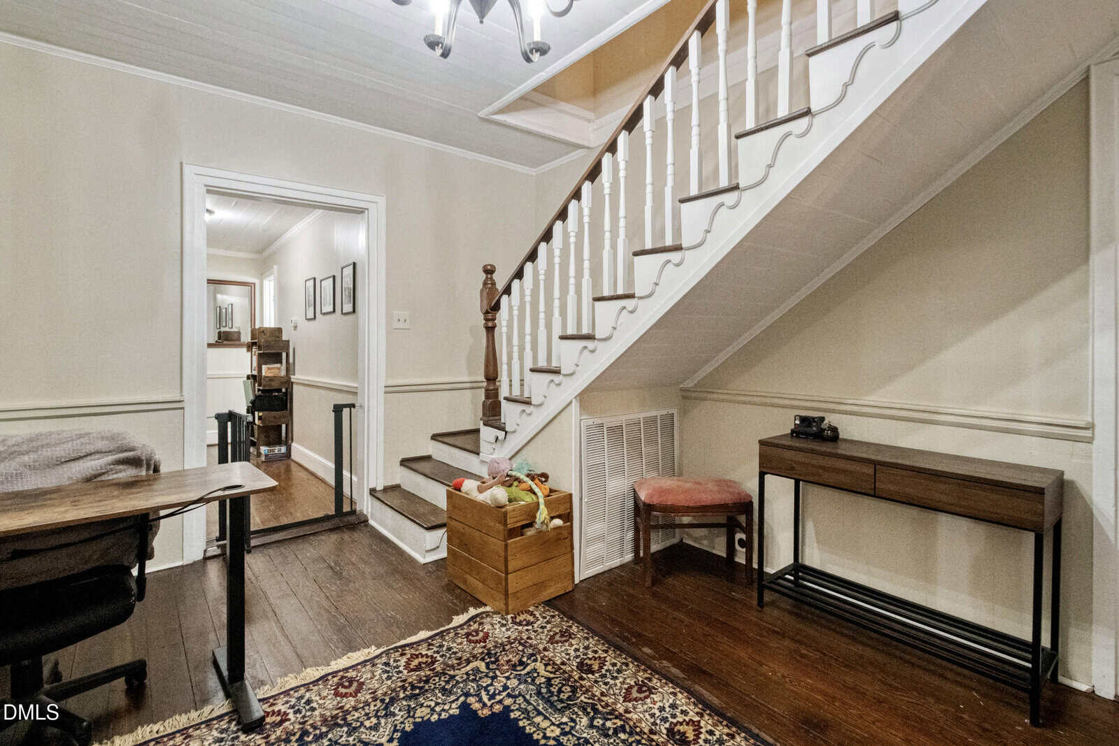 A room with dark wood floors, a patterned rug, a wooden staircase with white spindles, a desk, a console table, a pink bench, storage bins, and a hallway leading to another room.