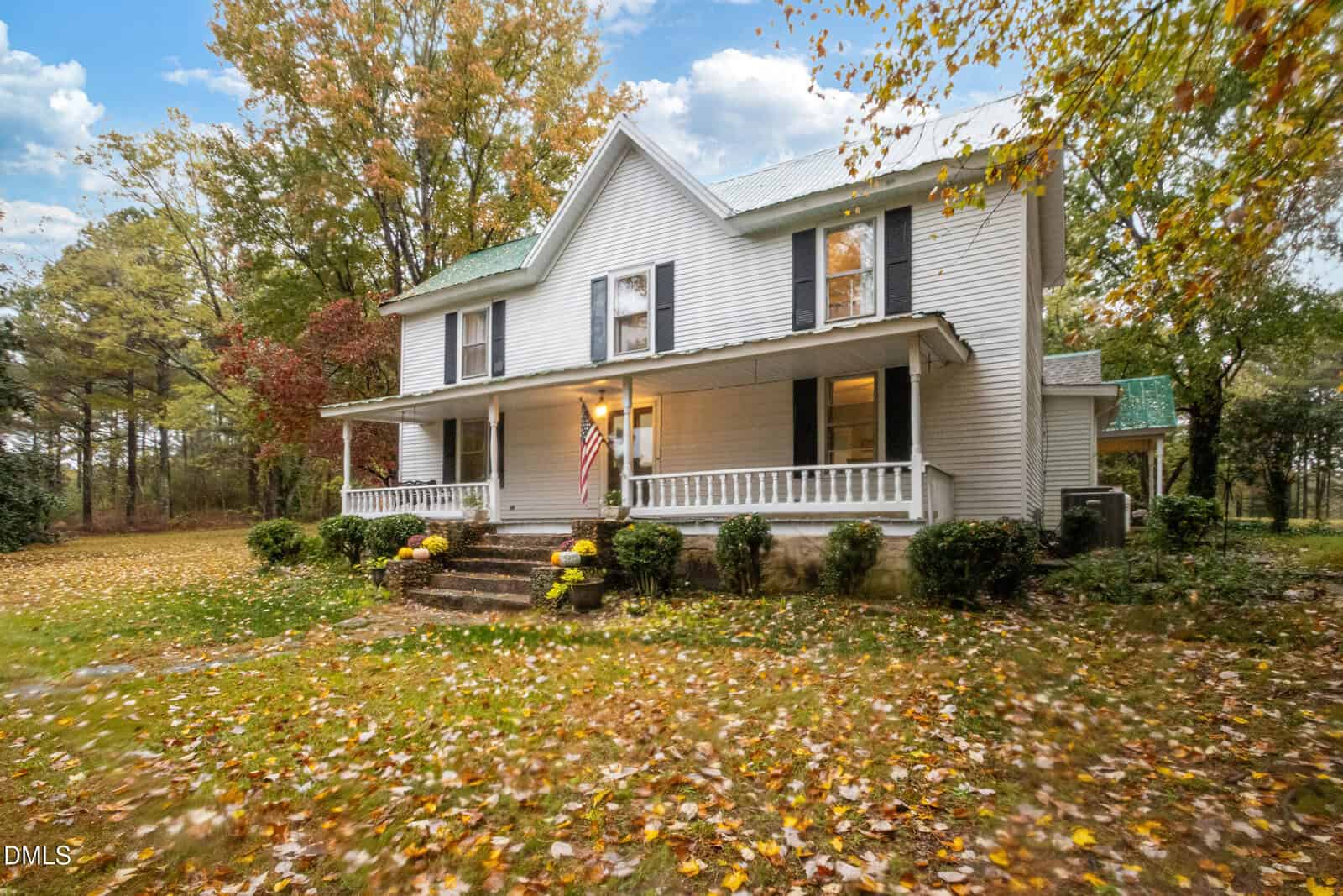 A two-story white house with a large front porch, green roof, and American flag. The yard is covered with autumn leaves and surrounded by trees. The house has shrubs and potted flowers along the porch steps.