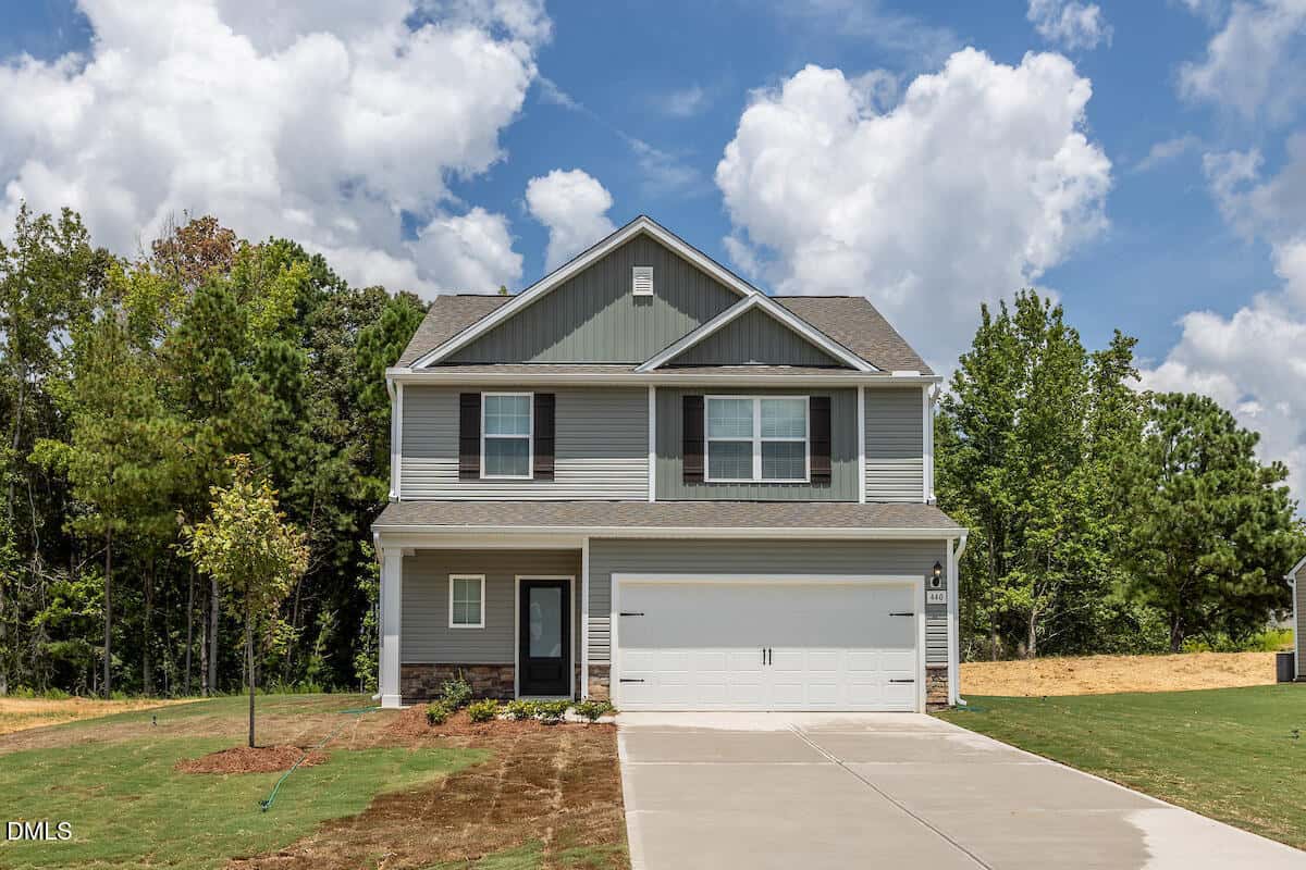 Two-story Oxford-style suburban house with gray siding, white trim, and a double garage at 1114 Rhino Bend. The home sits on a newly landscaped lawn with a young tree, bordered by woods and a partly cloudy sky above.