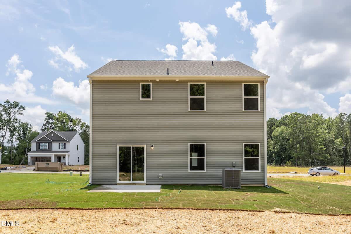 A two-story house with light gray siding, five rectangular windows, a sliding glass door, an exterior air conditioning unit, and a small concrete patio, with another house and trees visible in the background.