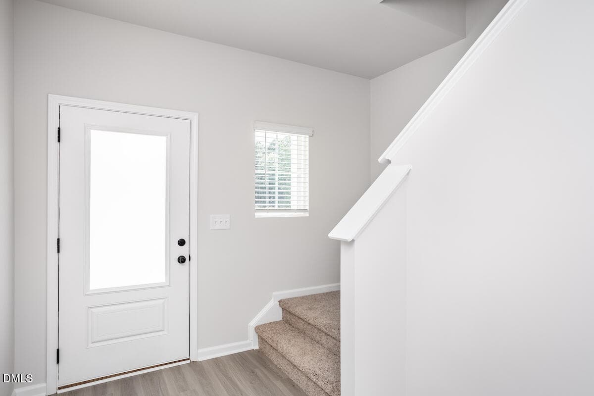 A clean, modern entryway with a white front door, a small window with blinds, a light switch, and carpeted stairs leading upward. The walls are painted light gray and the floor has light wood-style flooring.