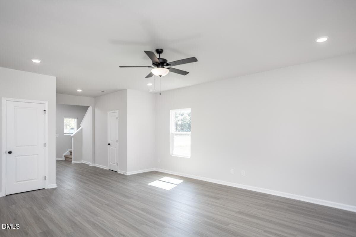 A bright, empty room with light gray wood flooring, white walls, a ceiling fan with a light, two white doors, one window, and a small stairway visible in the background.