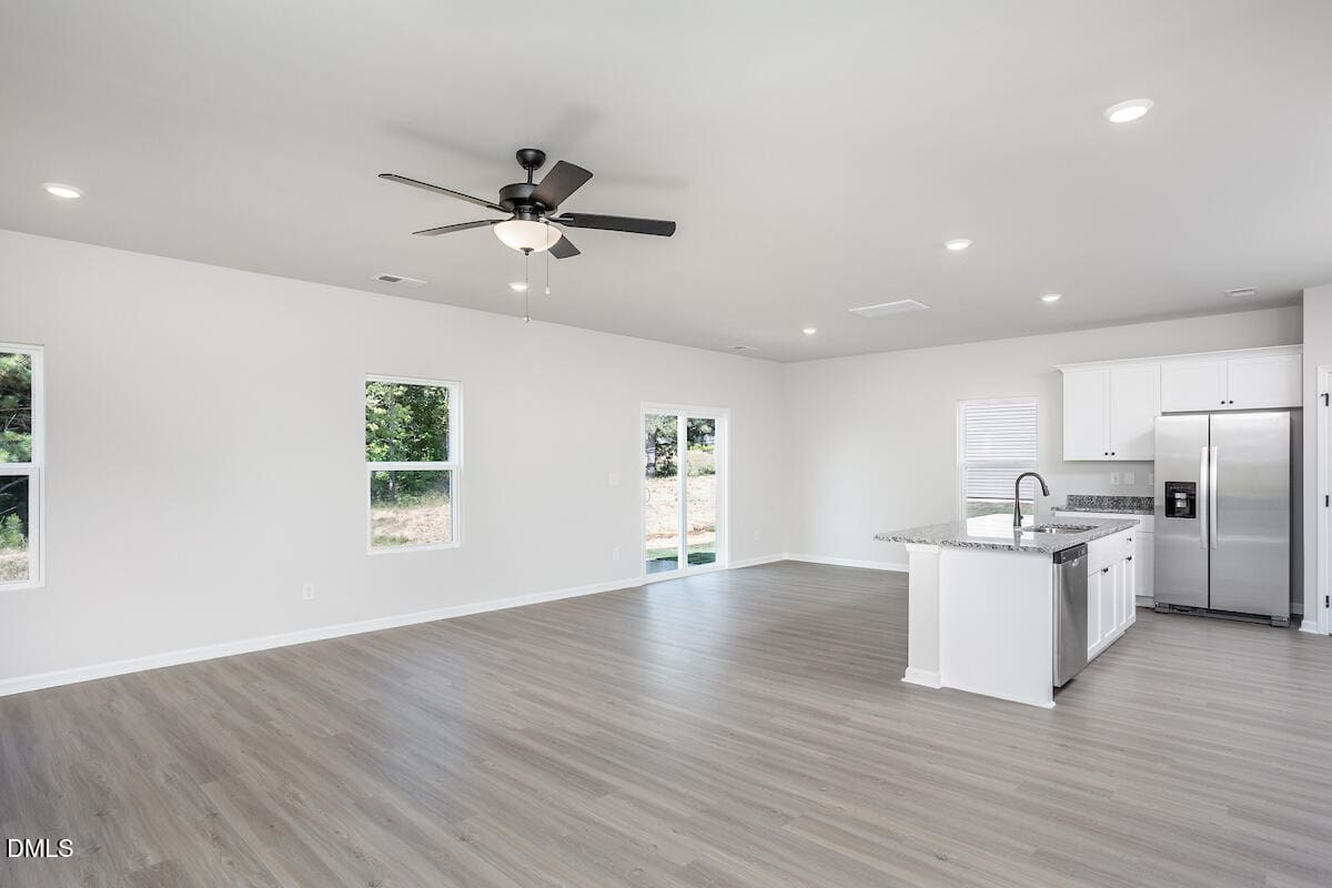A spacious, empty open-plan room with light wood flooring, white walls, and ceiling lights. The area includes a kitchen with white cabinets, granite countertops, and stainless steel appliances. There are multiple windows and a ceiling fan.