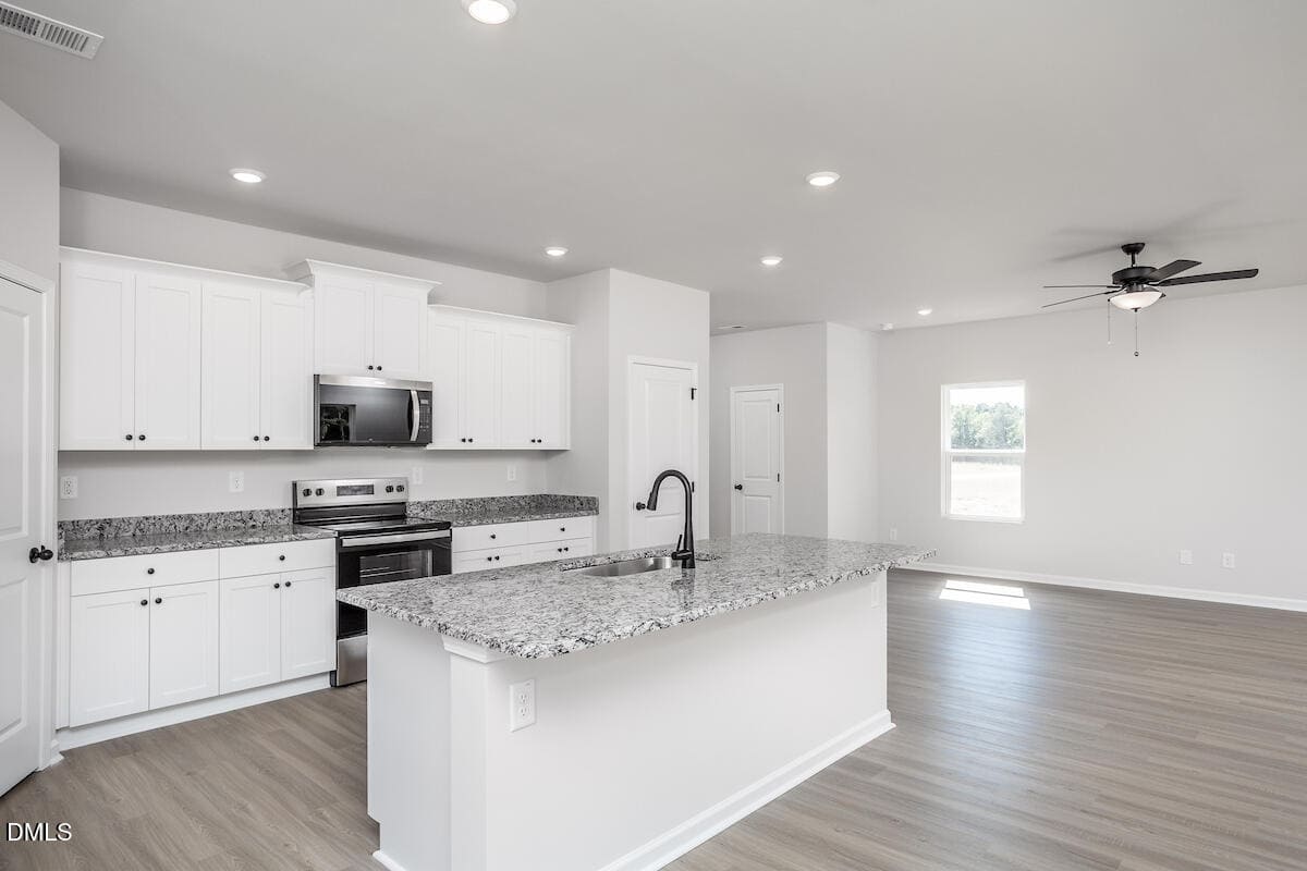 A modern kitchen with white cabinets, stainless steel appliances, a gray granite island with a black faucet, light wood flooring, and recessed ceiling lights, leading to a bright open living area with a ceiling fan.