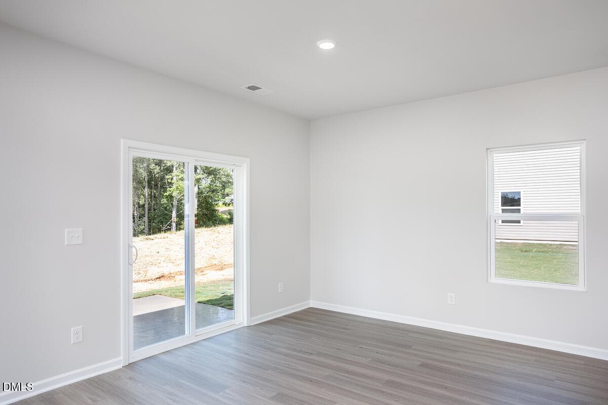 A bright, empty room with light gray walls and wood-style flooring features a sliding glass door leading outside and a single window. The space is well-lit with natural light.