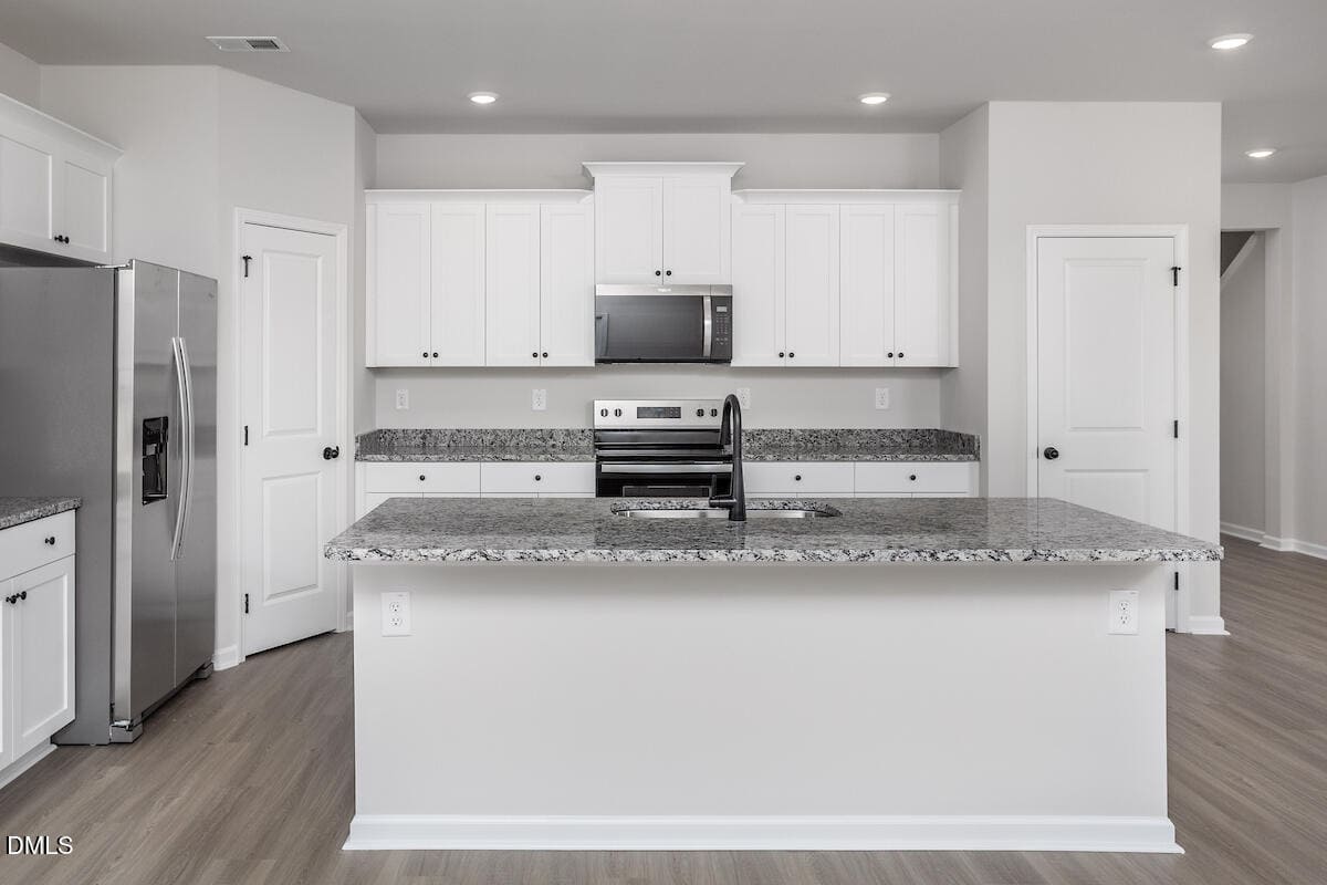 A modern kitchen with white cabinets, granite countertops, stainless steel appliances, a center island with a sink, and light wood flooring. The walls are painted light gray and recessed lighting is installed in the ceiling.