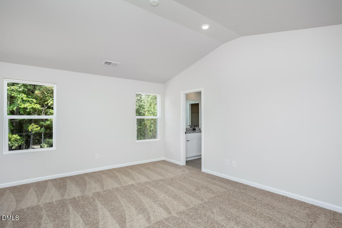 A clean, empty room with light-colored walls, two windows showing greenery outside, beige carpet with vacuum lines, and an open doorway leading to a bathroom with white cabinets.