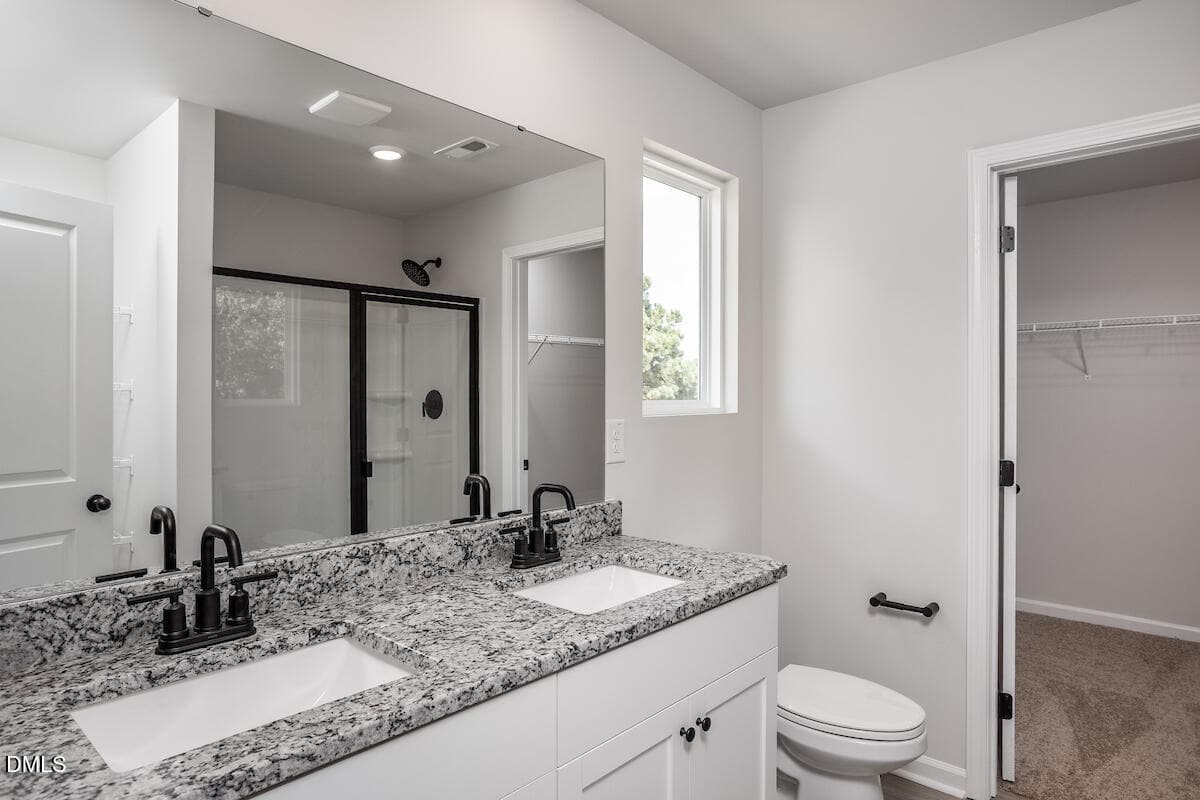 Modern bathroom with double sinks, granite countertop, black fixtures, large mirror, toilet, glass shower, window, and an open walk-in closet with shelving. White walls and neutral-toned flooring complete the space.