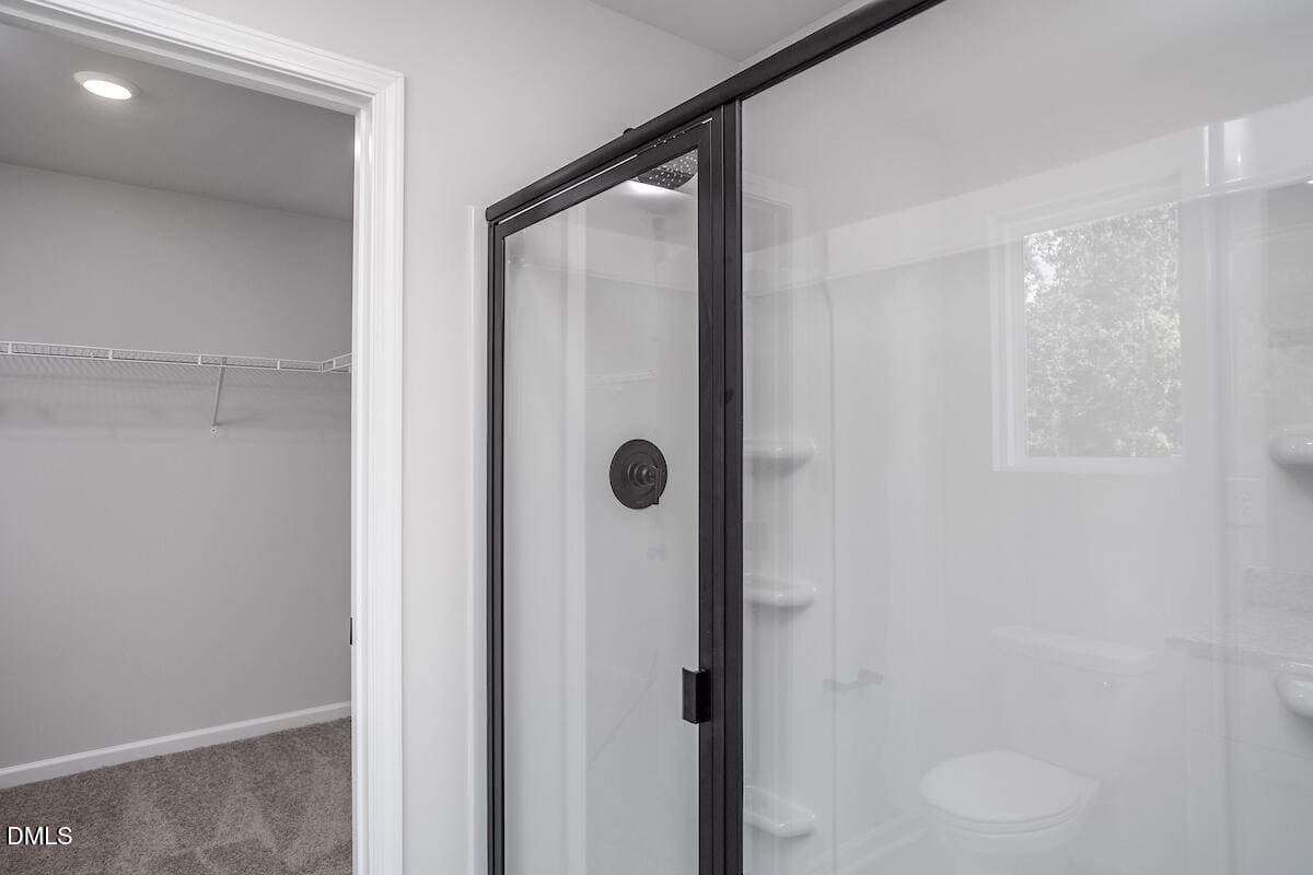 A modern bathroom with a glass shower enclosure, black trim, built-in shelves, and a window. Part of a carpeted walk-in closet with wire shelving is visible to the left.
